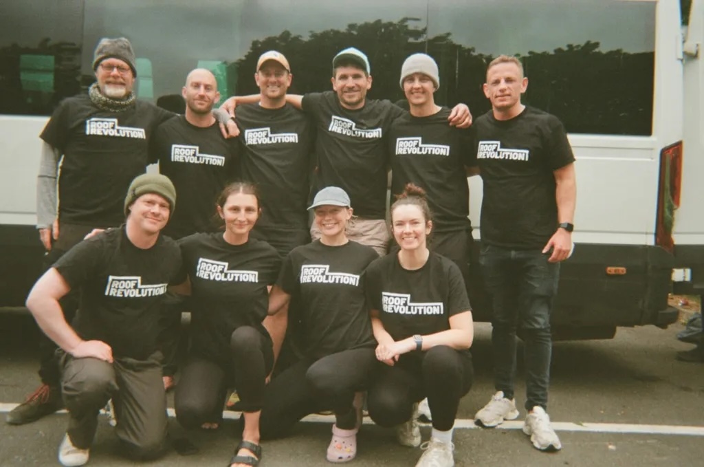 Ten people wearing matching black t-shirts posing in two rows, six standing behind and four seated in front, next to a white vehicle.