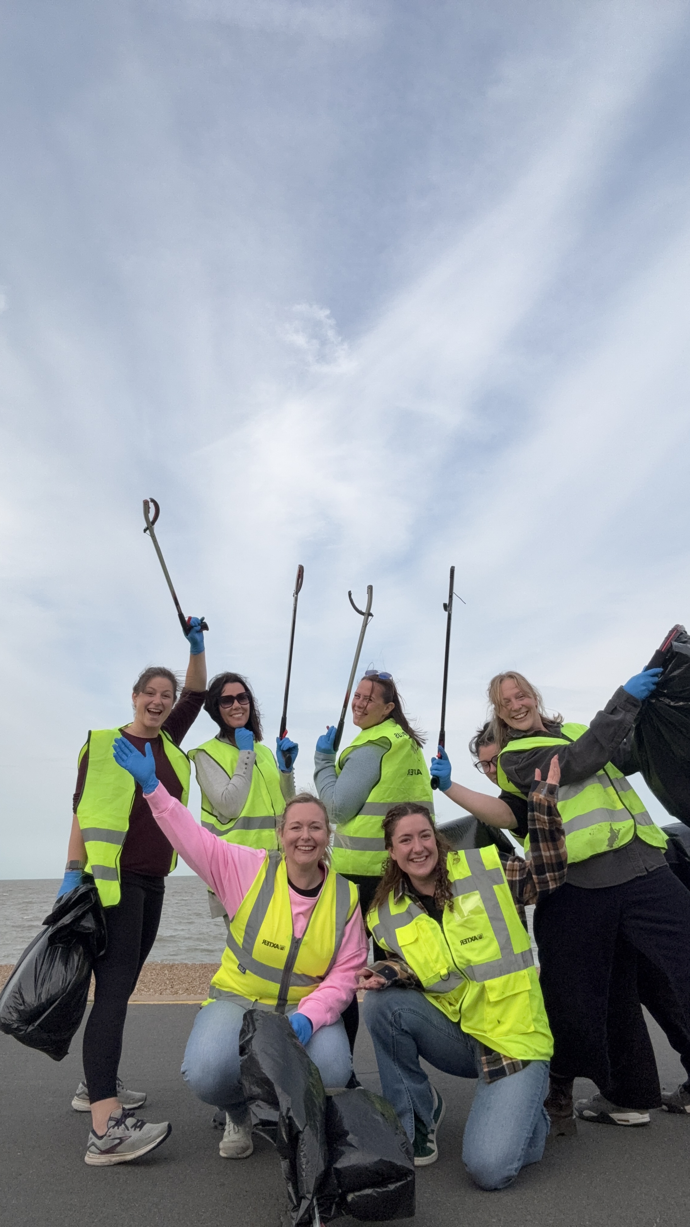 Group of seven women in bright yellow hi-vis vests holding litter-picking sticks and rubbish bags on a beach under cloudy sky.