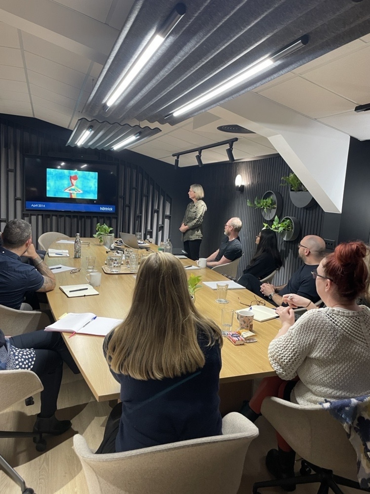 Business meeting in modern conference room with wooden table, white chairs, wall-mounted TV screen, and presenter standing beside seated attendees.