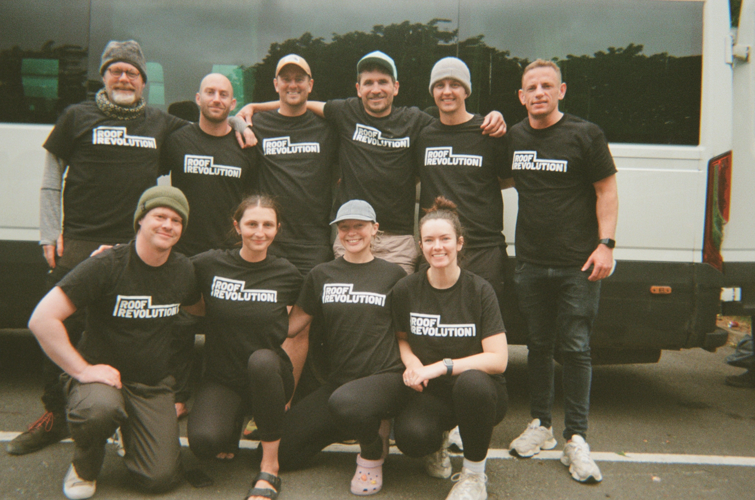 Ten people wearing matching black "Rock Revolution" t-shirts pose in two rows in front of a white tour bus.