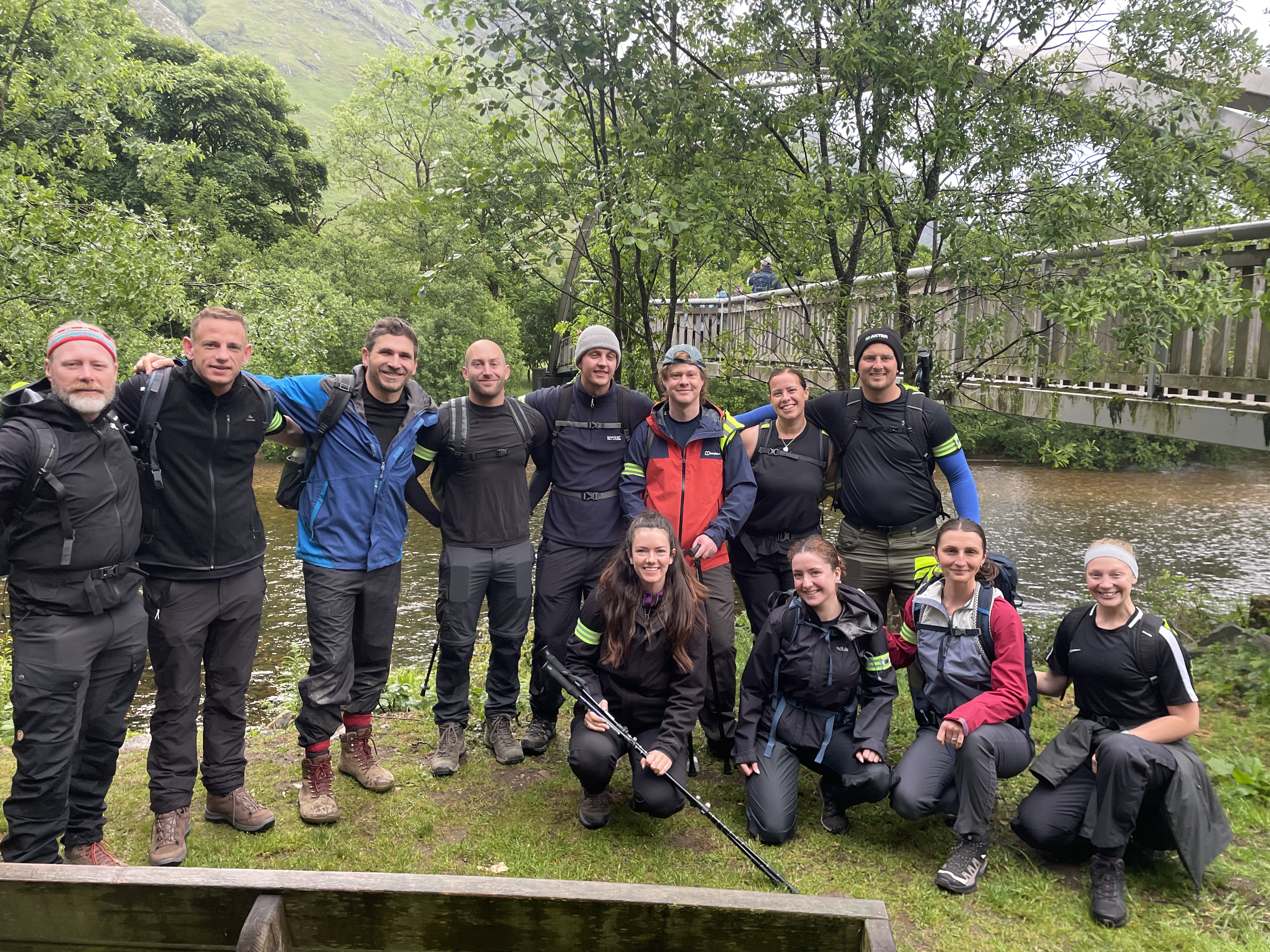 Group of thirteen people in outdoor gear posing by river with bridge and green hills behind them.