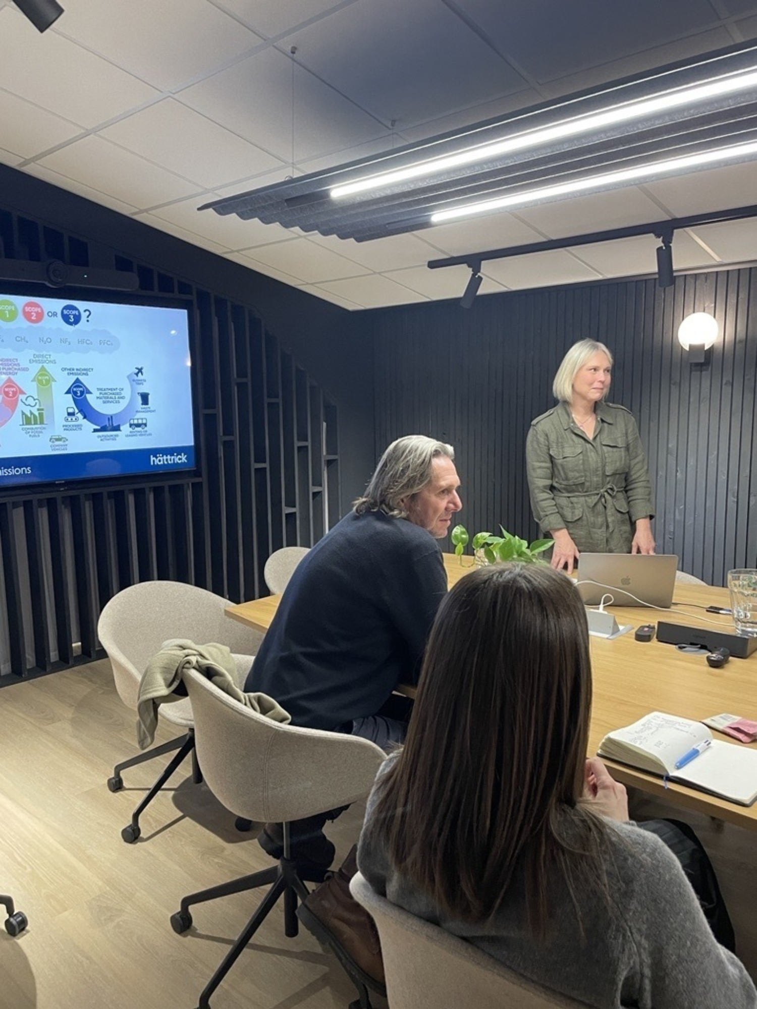 Conference room with wooden table, beige chairs, and wall-mounted screen. Three people present - woman standing, man seated at table, woman in foreground.