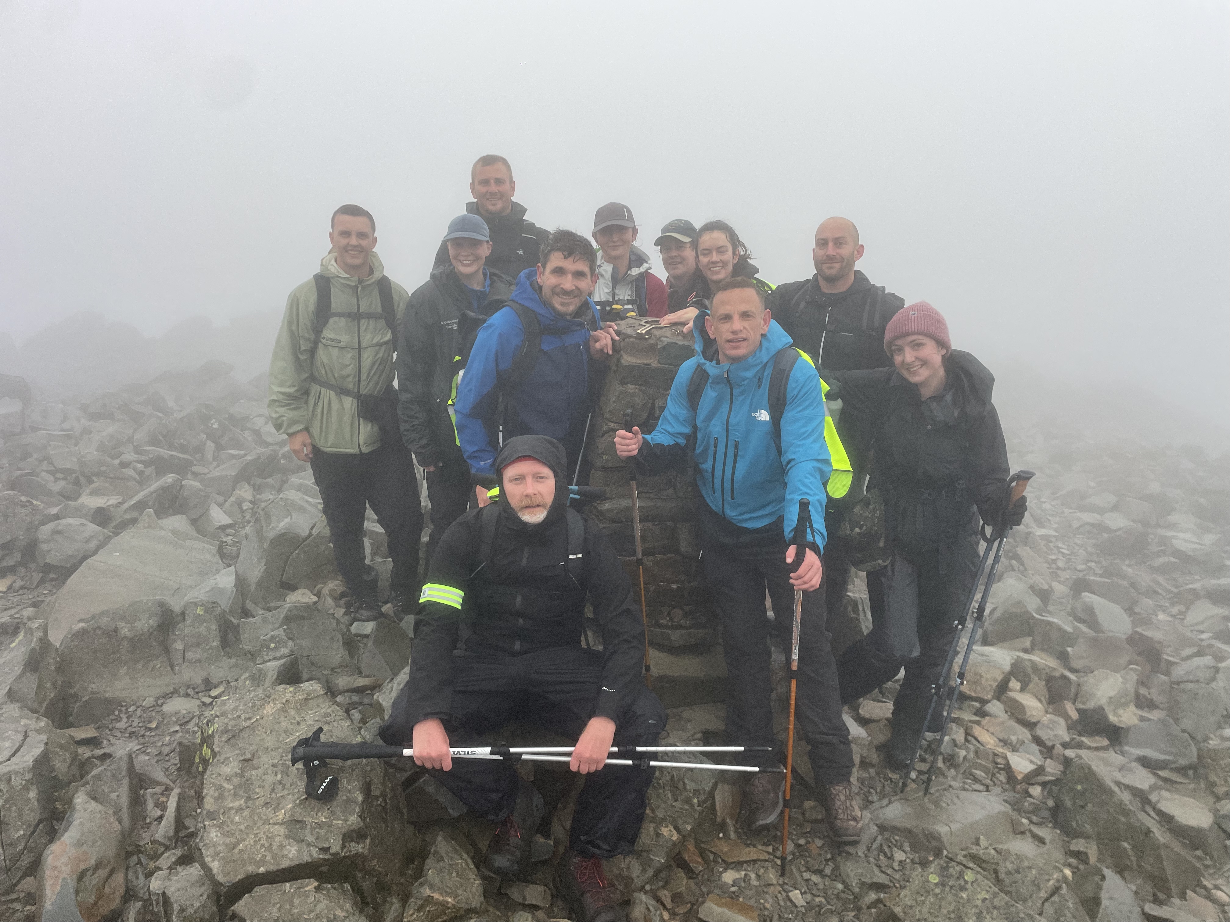 Group of hikers in waterproof jackets and woolly hats gathered on rocky mountain summit in thick fog and mist.