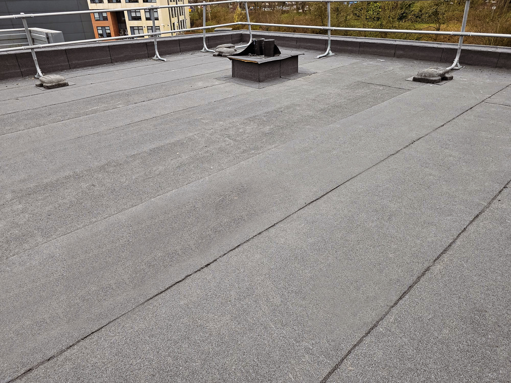 Flat rooftop with grey felt membrane, a central metal vent, two small roof lights, metal railings, and a brick building visible in the background.