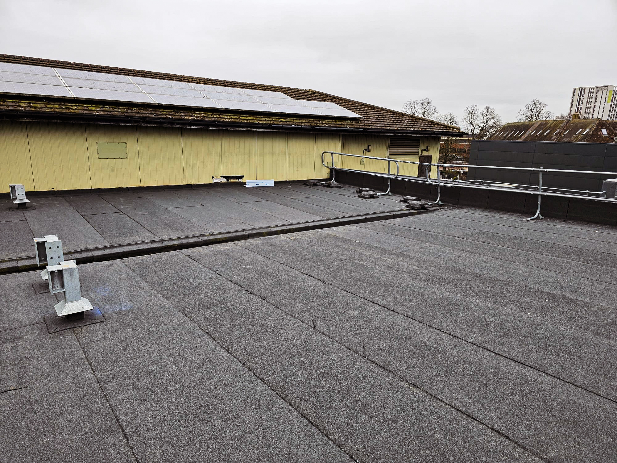 Flat rooftop with dark grey felt surface, metal safety railings, cable, and mounting brackets. Solar panels on adjacent pitched roof. Overcast sky.