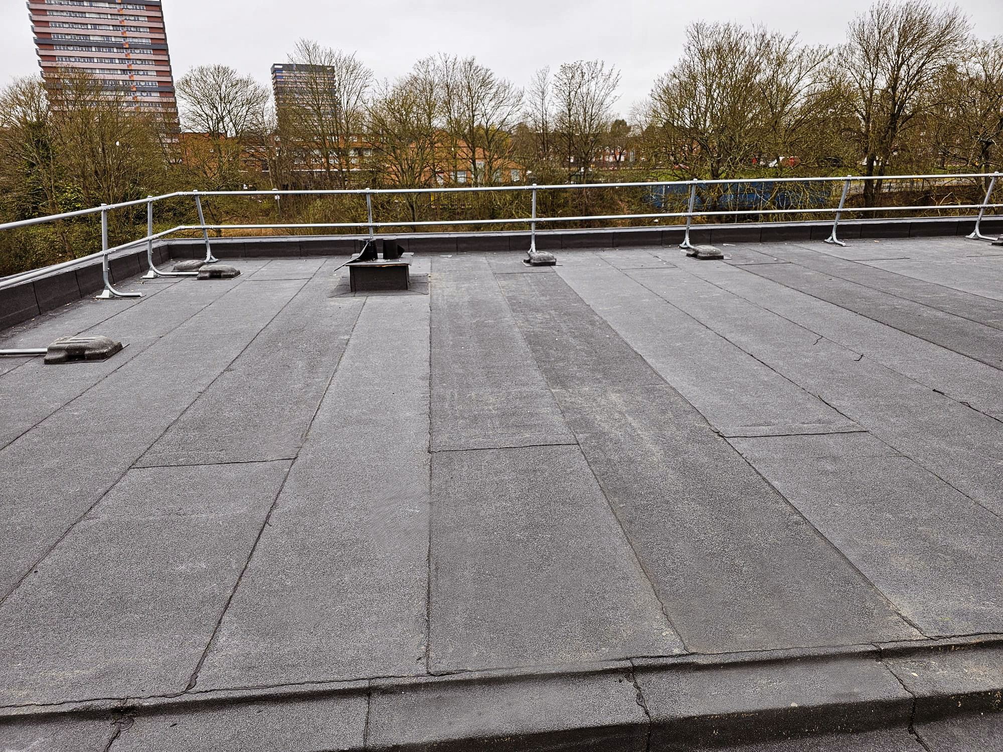 Flat rooftop with grey felt surface, white safety railings, rooftop fixtures, and bare trees with tower blocks visible in the background.