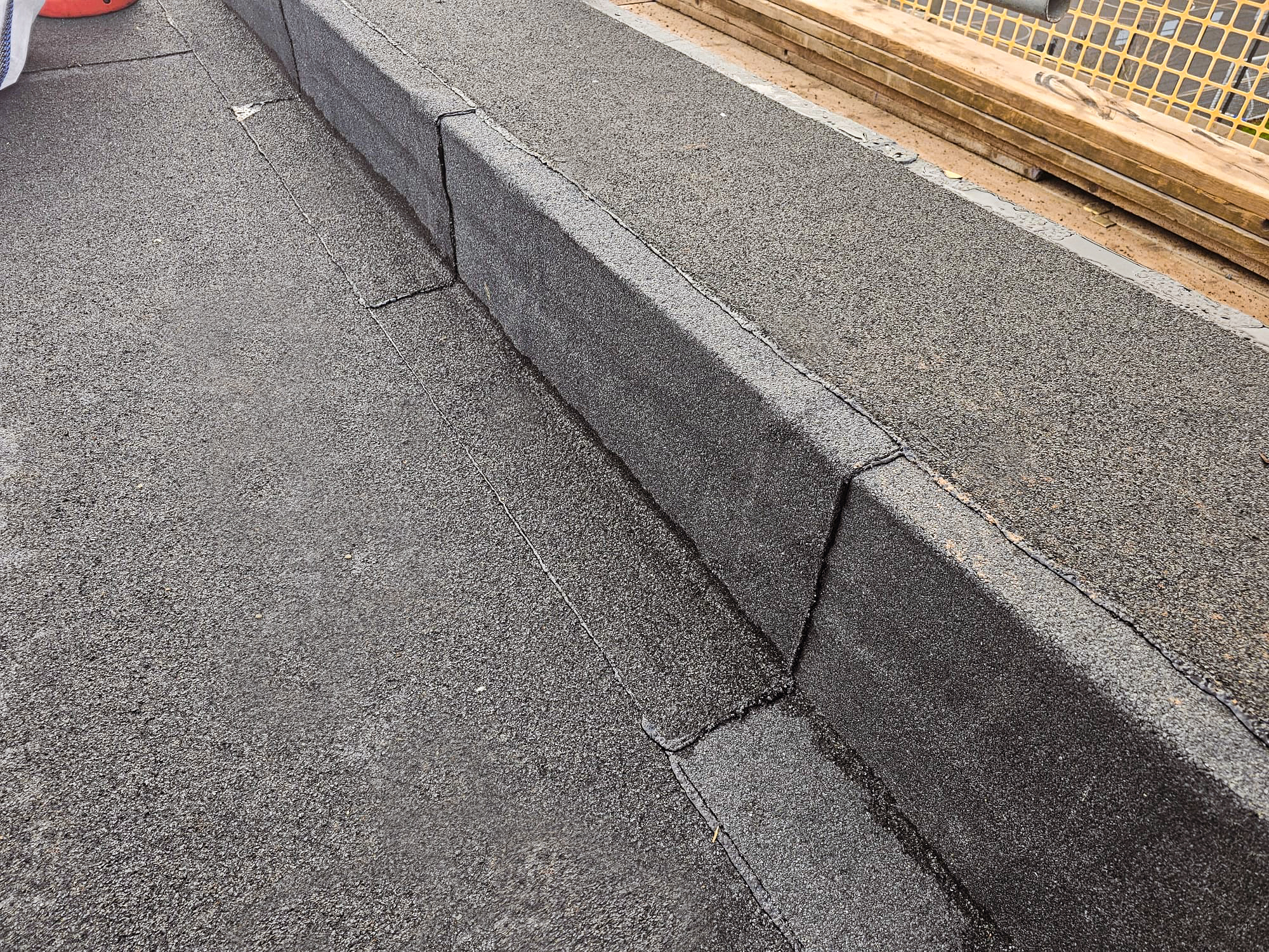 Dark grey granite kerb stones forming a raised edge between two levels of aggregate surface, with timber and metal fencing visible in background.