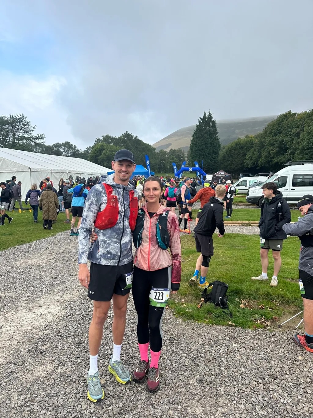 Two runners with race numbers and medals at outdoor event finish area. White marquee, hills, trees in background. Cloudy sky.