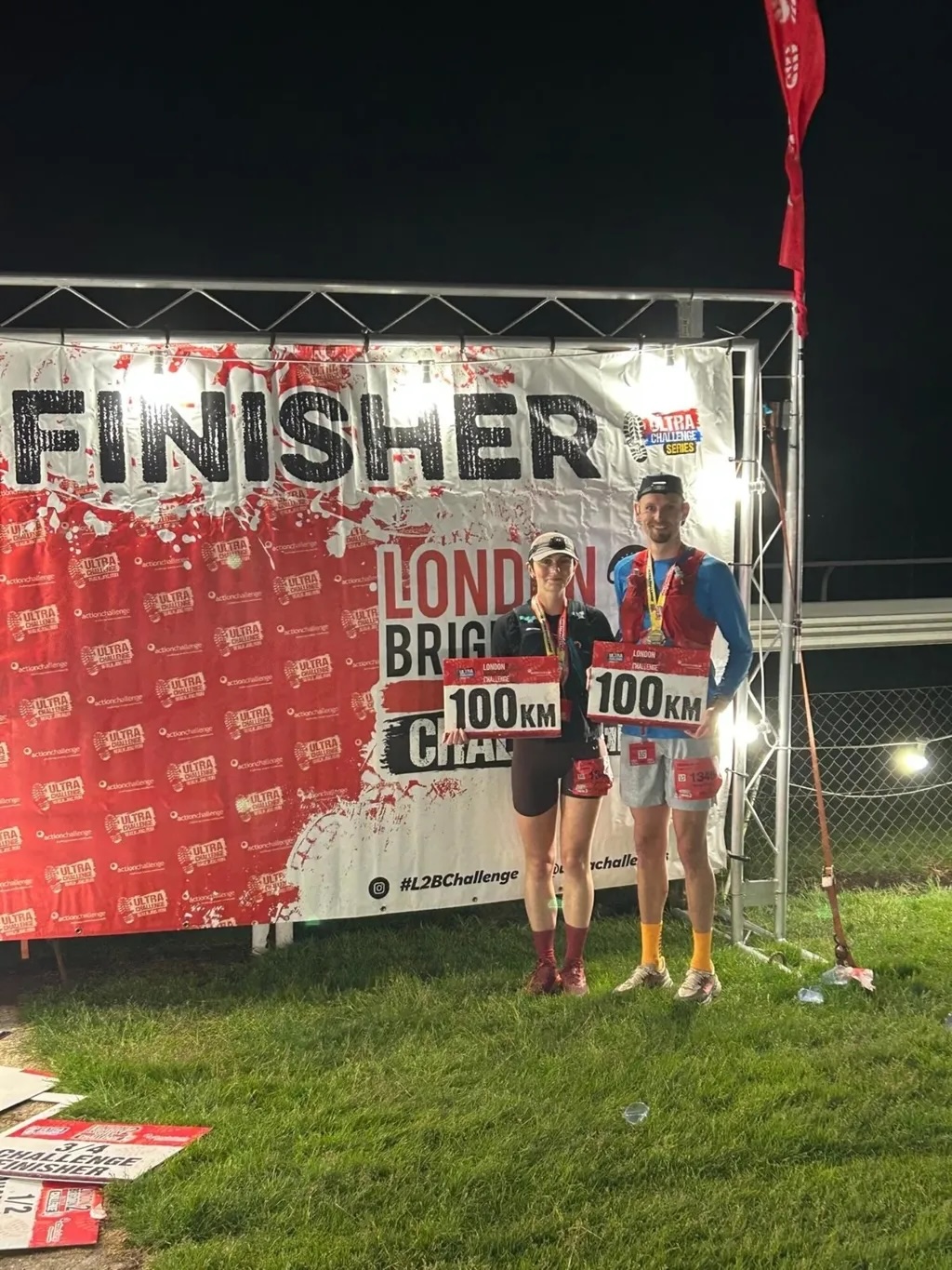 Two runners with medals posing at night-time race finish line with "FINISHER" banner and "LONDON BRIGHTON 100KM" signage.