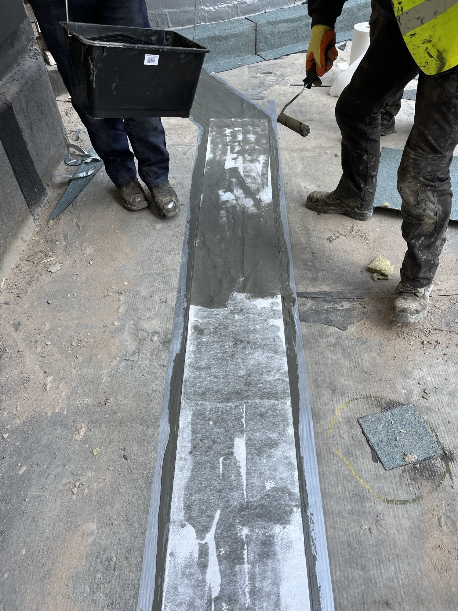 Workers in boots standing around clear glass channel or trough on concrete floor, with black bucket and tools visible.