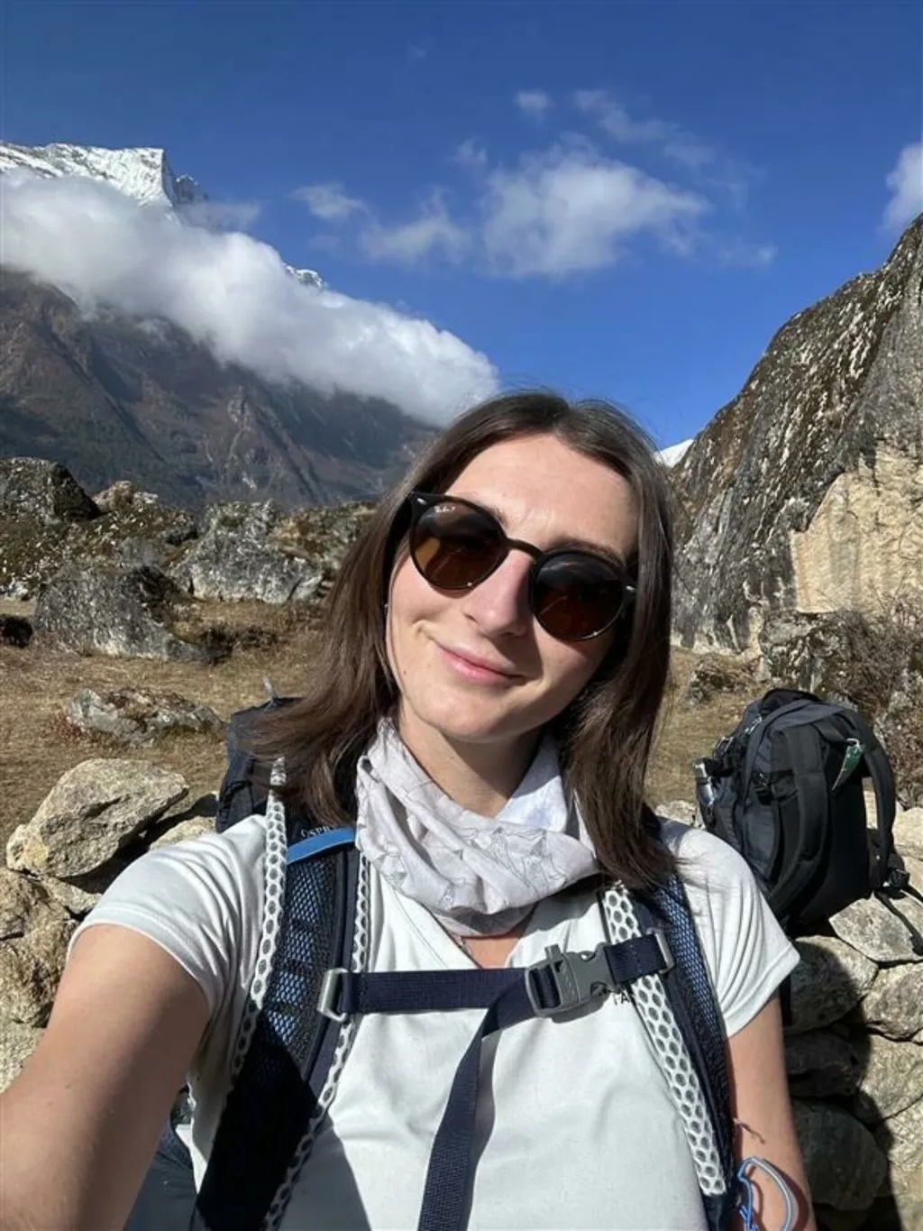 Woman in sunglasses and hiking gear taking selfie in mountainous terrain with snow-capped peaks and blue sky with white clouds.