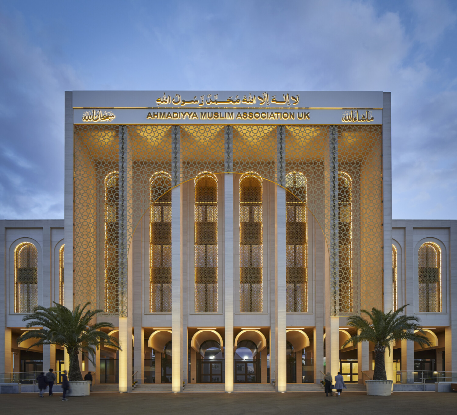 Large white building with Islamic architectural features, golden geometric patterns, arched windows, and illuminated entrance with palm trees.