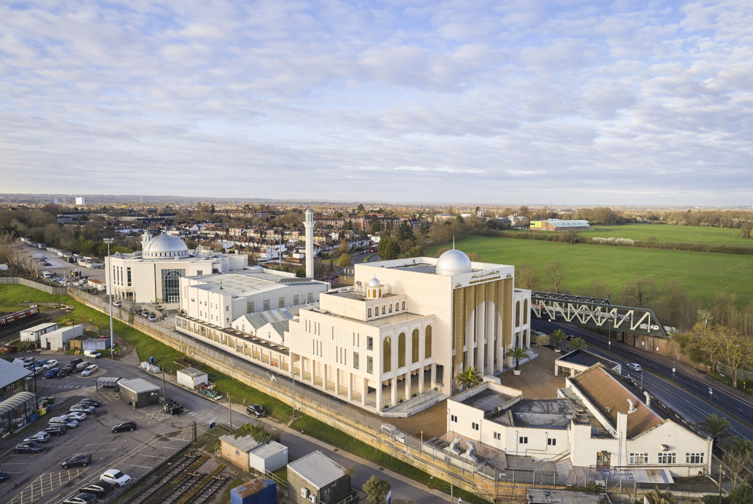 Aerial view of large white mosque with minaret and dome in suburban setting, surrounded by car parks, roads, and green fields.