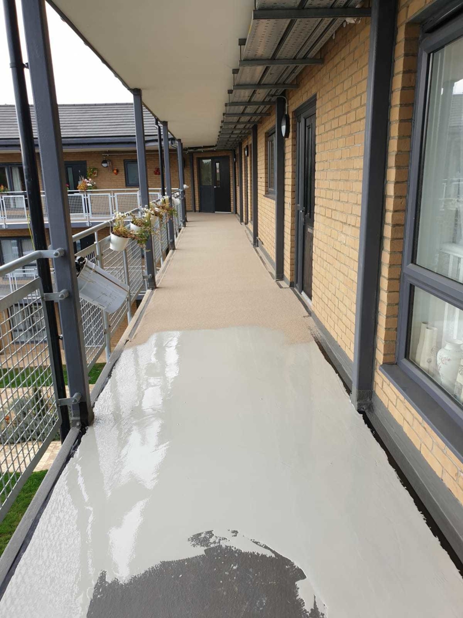 External concrete walkway with metal railings alongside brick building with black window frames and doors.