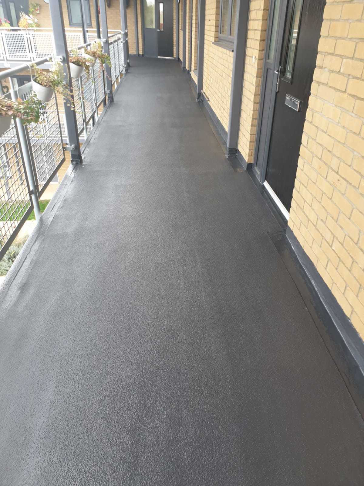 External corridor with grey flooring, cream brick walls, metal railings on left, and dark doors along right side.