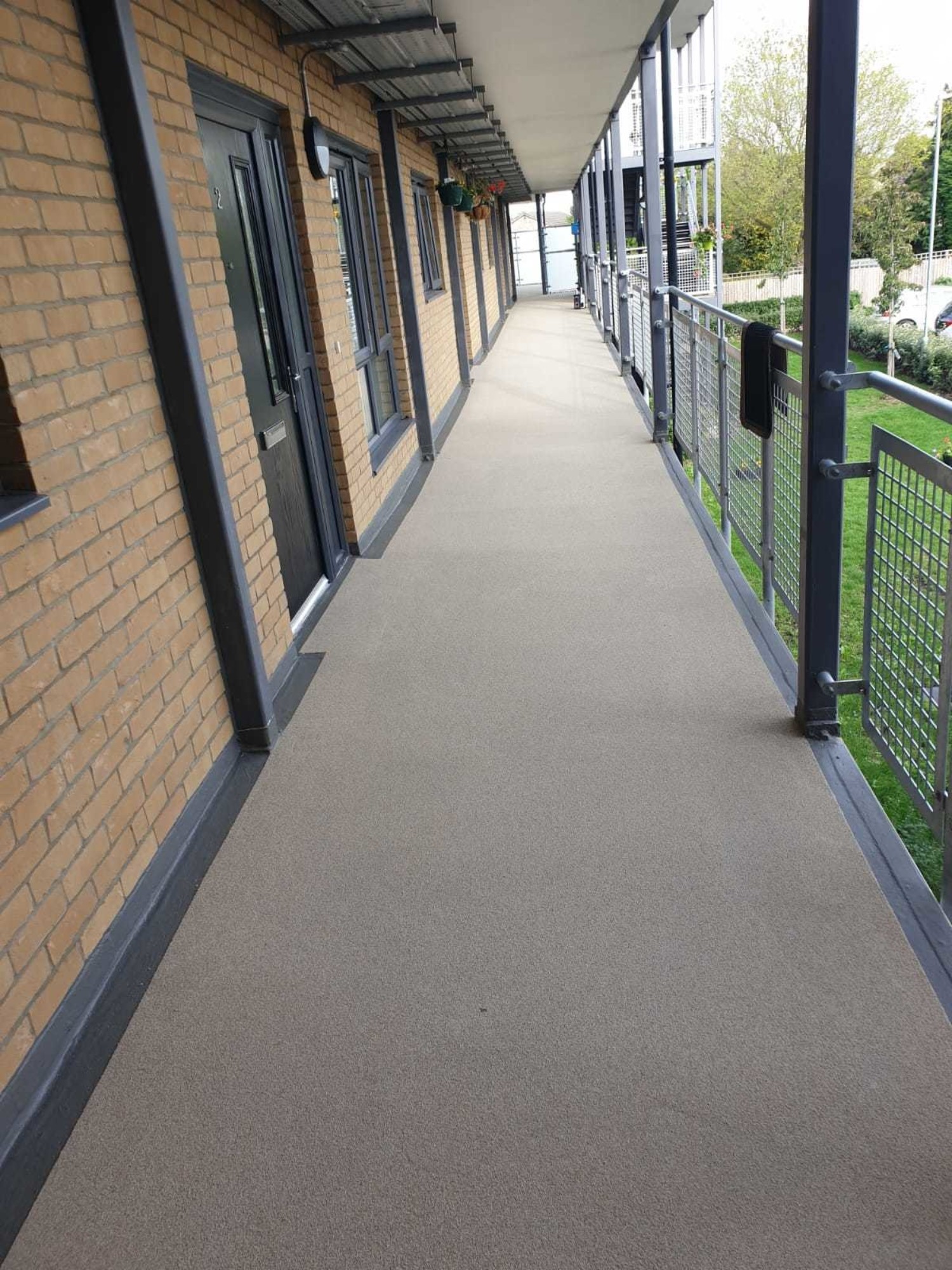 External walkway with dark doors along brick wall, metal railings on right overlooking green lawn area below.
