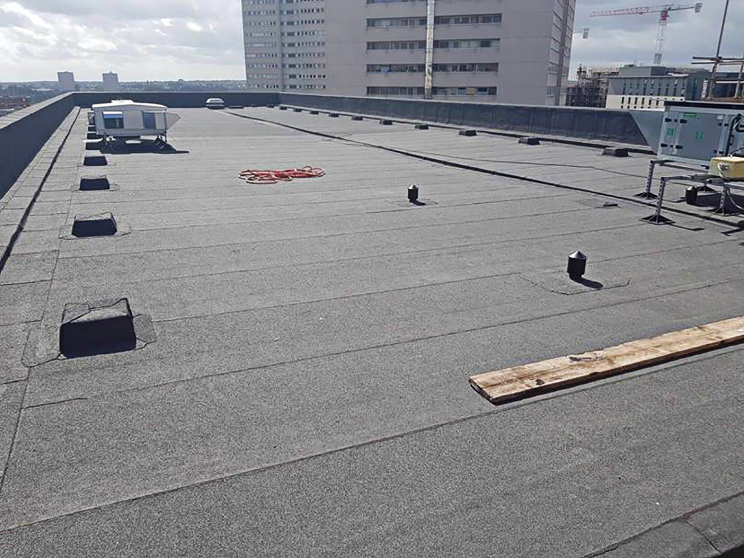 Flat rooftop with grey membrane, red circular marking, ventilation units, and high-rise buildings in background under cloudy sky.