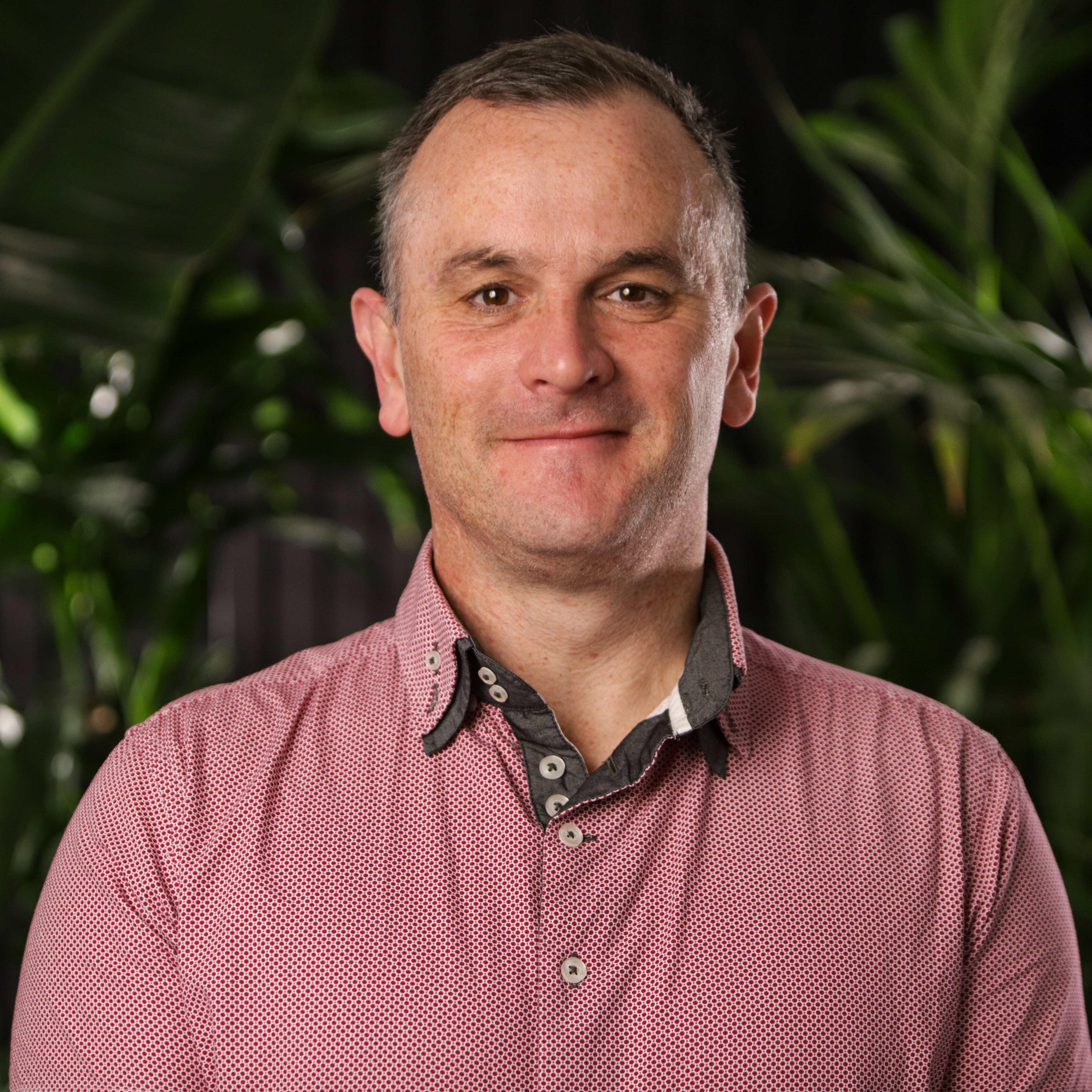 Middle-aged man with short grey hair wearing red patterned shirt, smiling at camera with green foliage in background.