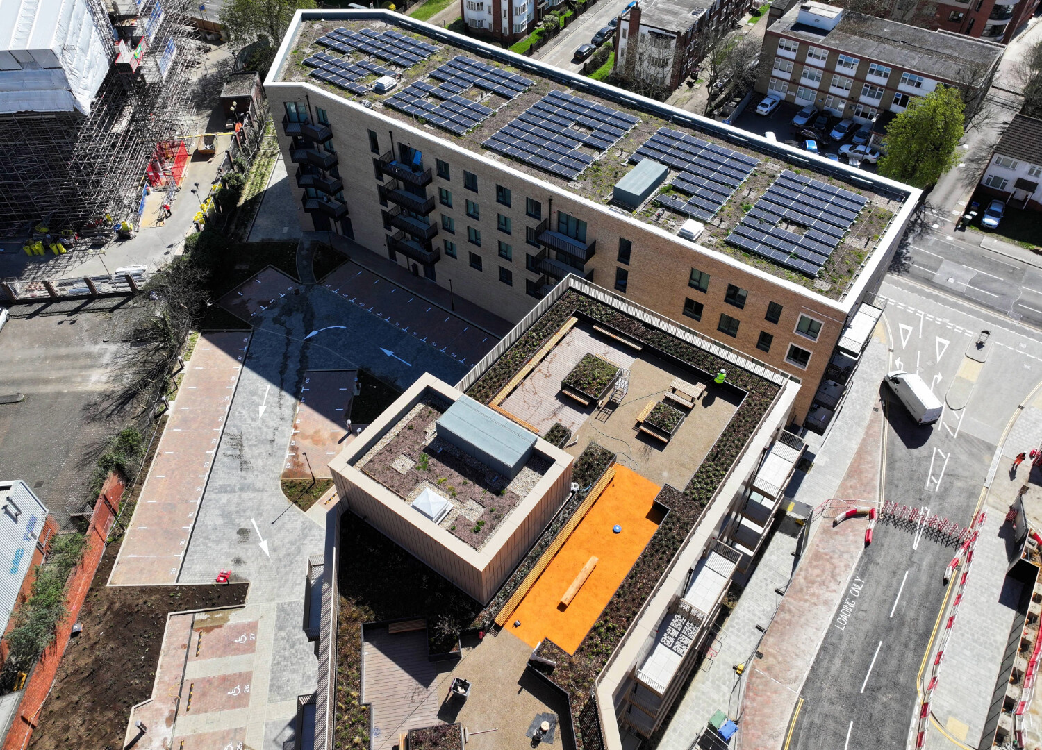 Aerial view of modern building with blue solar panels on roof, orange courtyard area, and surrounding urban development.