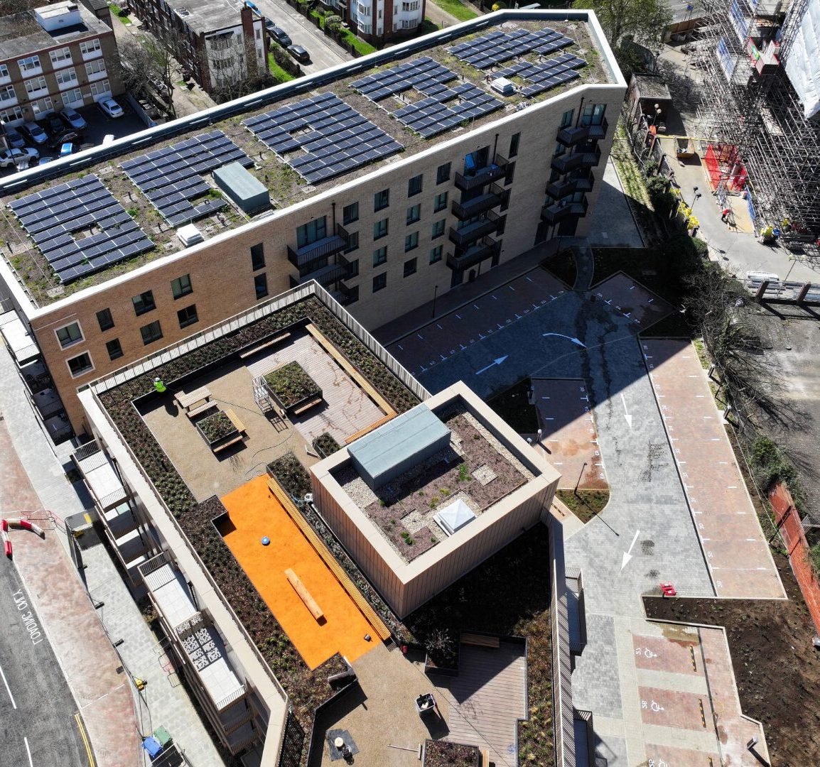 Aerial view of modern building with blue solar panels on flat roof, orange courtyard area, and surrounding urban streets.