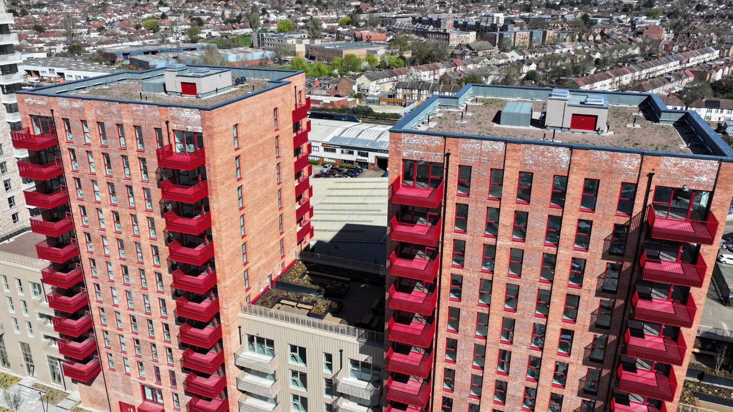 Two tall residential tower blocks with red brick facades and bright red balconies, aerial view showing surrounding urban landscape.