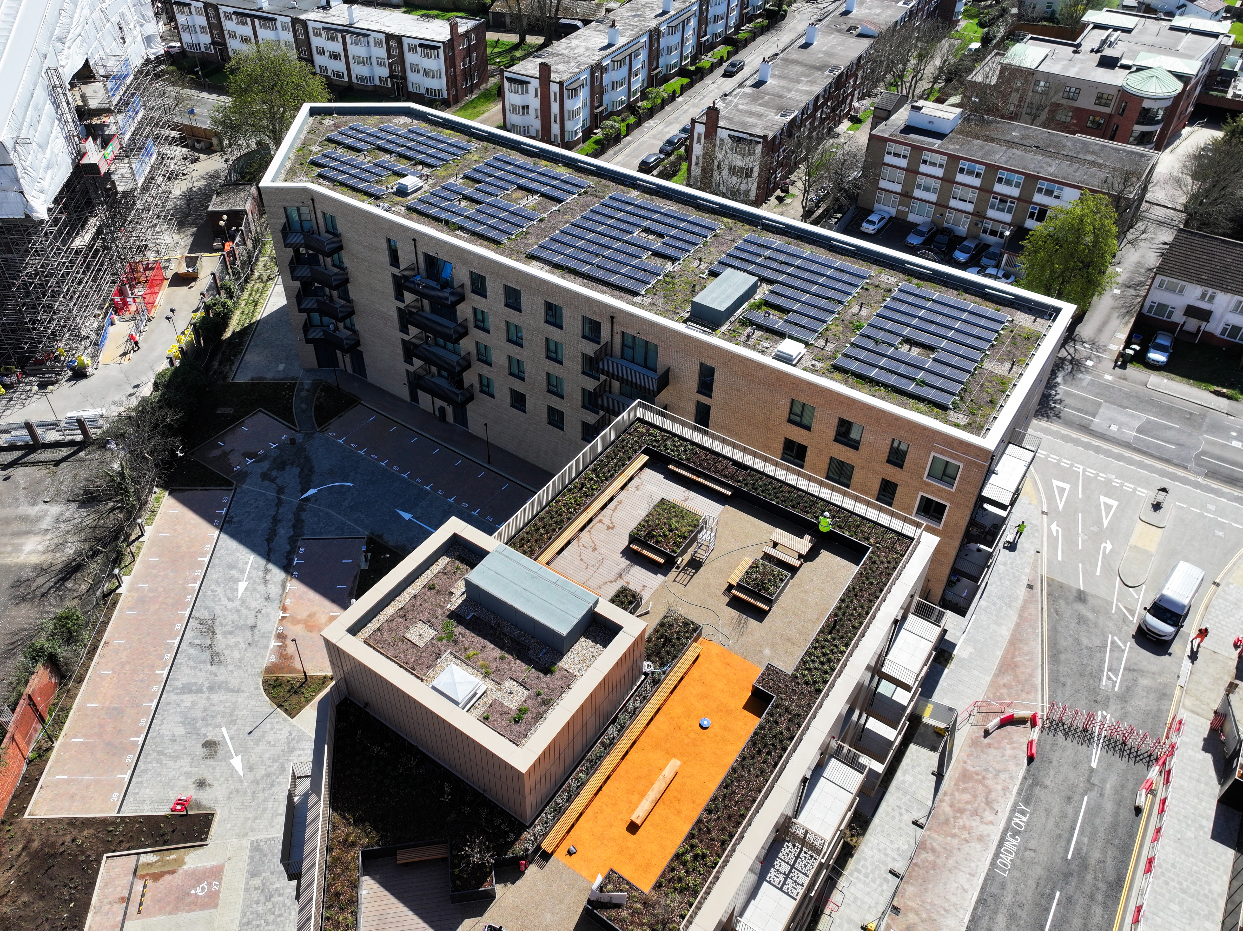 Aerial view of modern residential building with solar panels on roof, orange courtyard feature, surrounded by urban streets and housing.