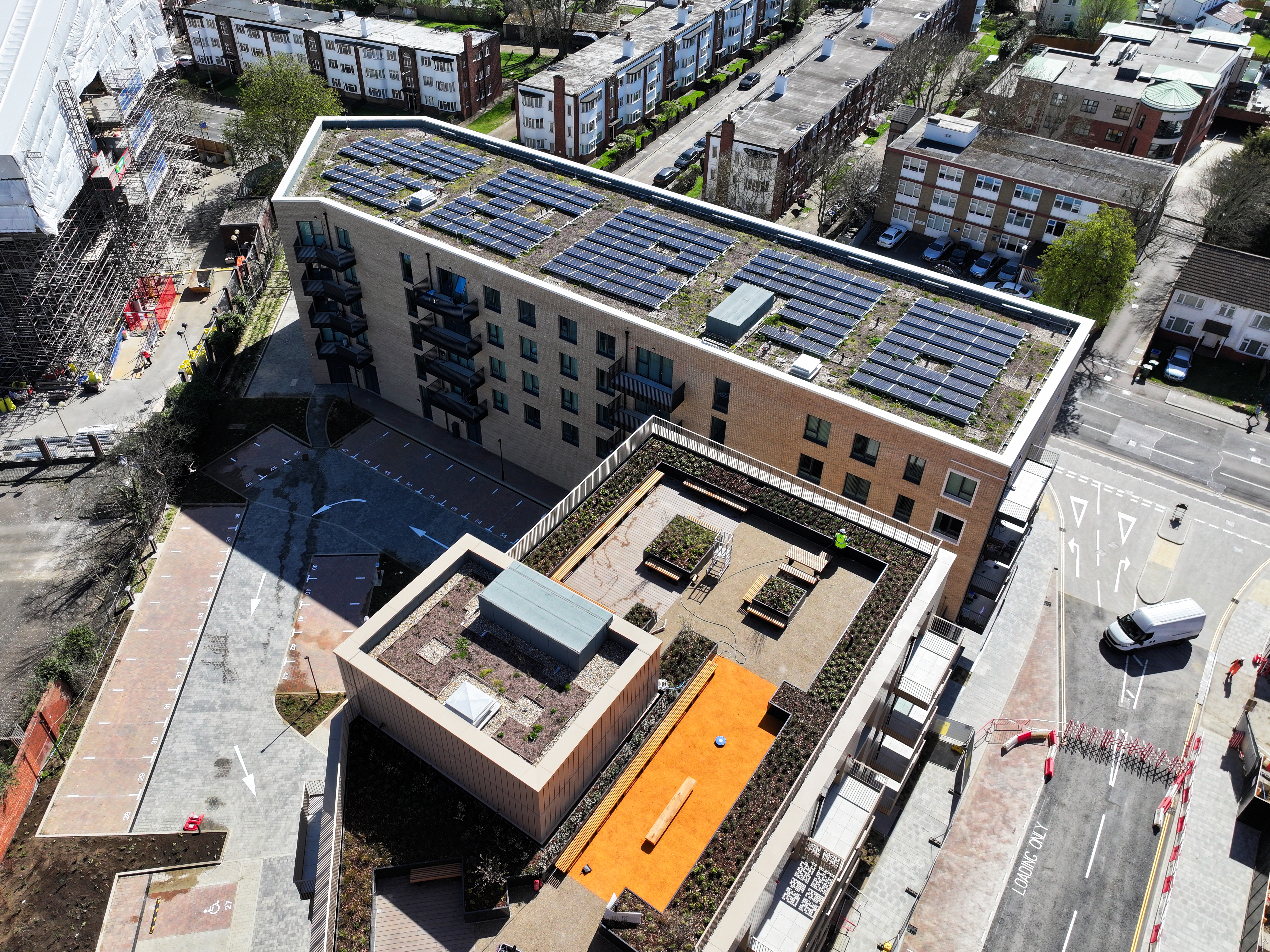 Aerial view of modern multi-storey building with solar panels on roof, orange courtyard area, surrounded by residential streets.