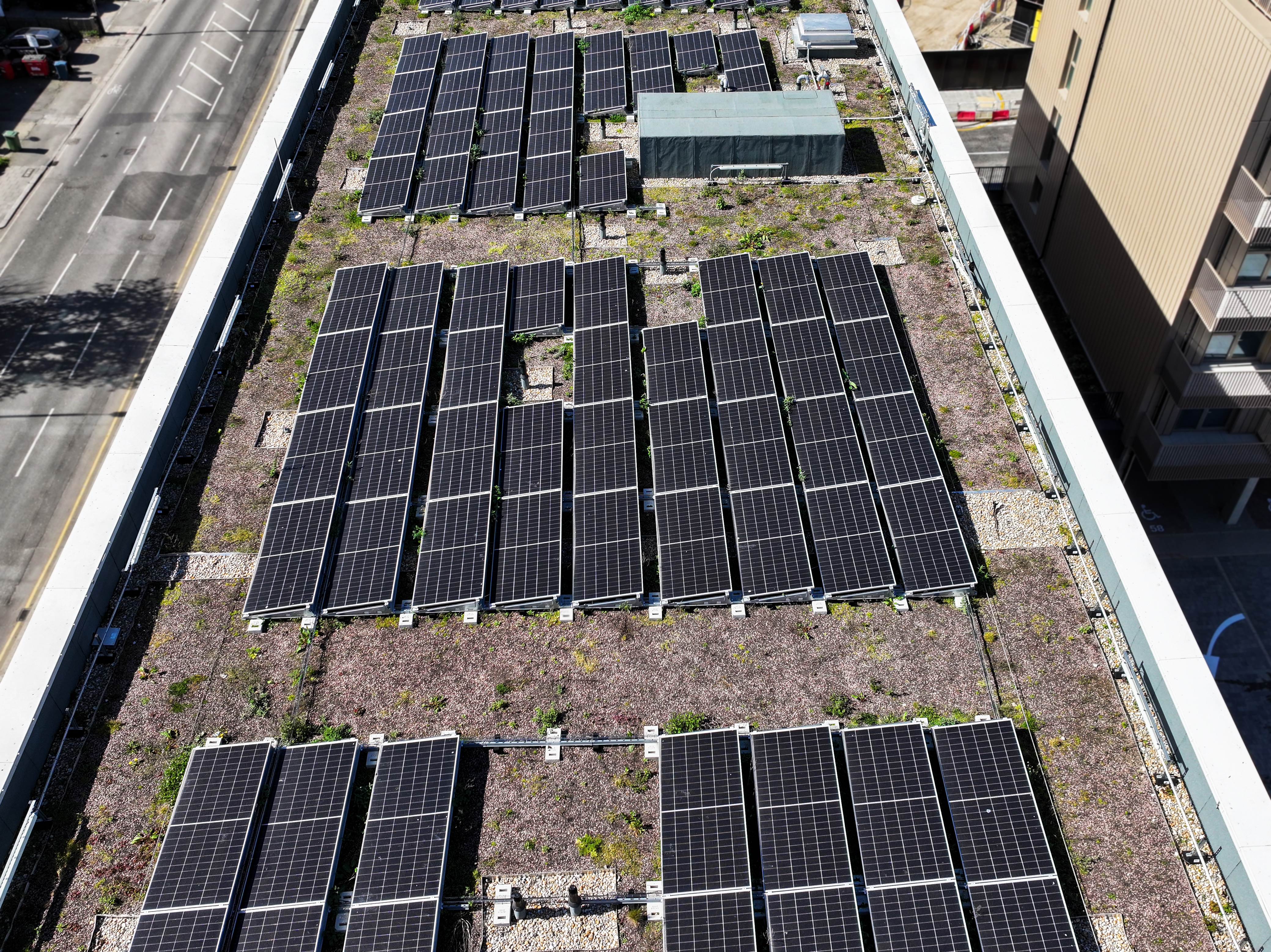 Aerial view of rooftop solar panel installation with multiple dark blue rectangular arrays arranged in rows on brown flat roof surface.