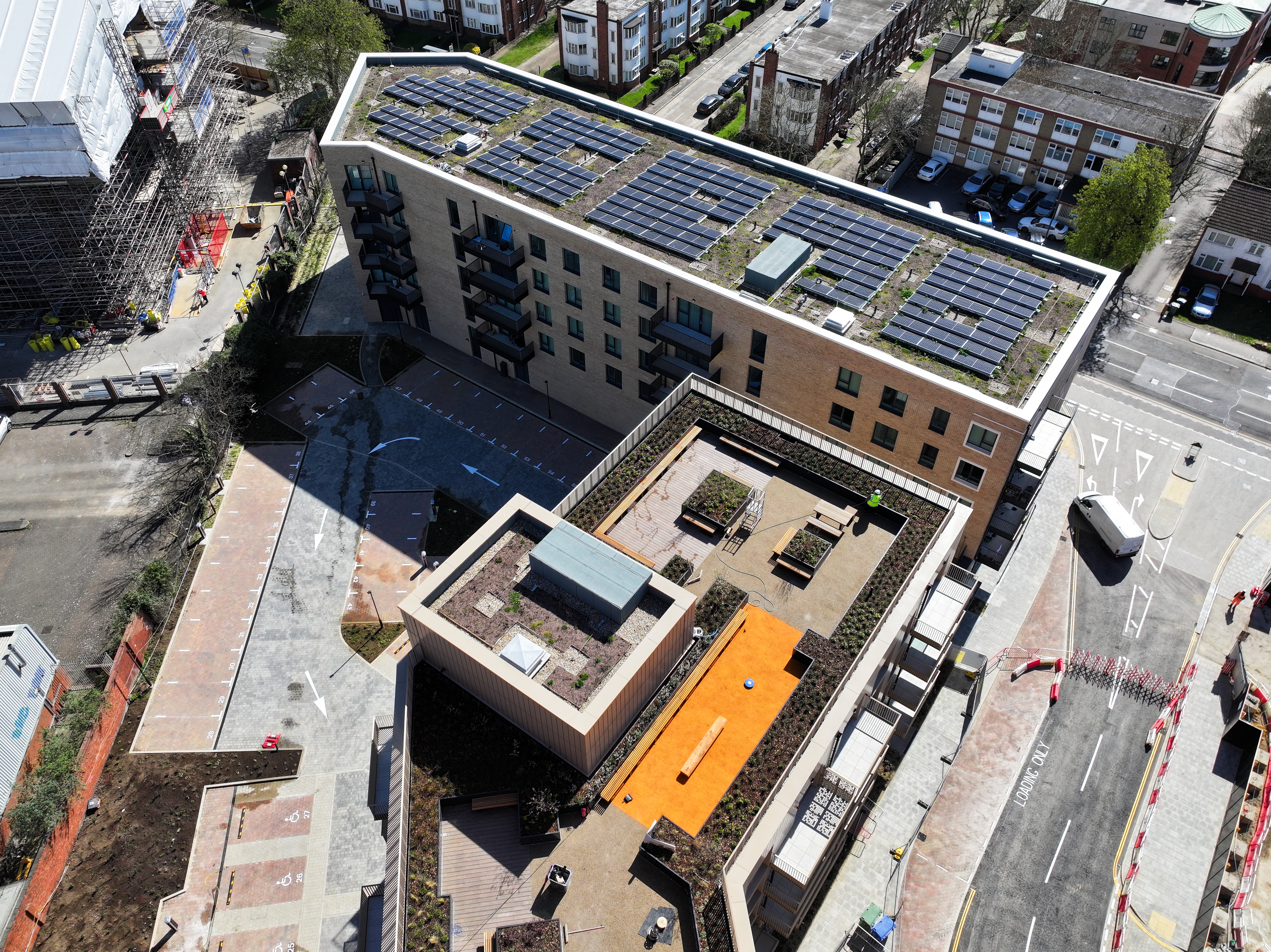 Aerial view of modern residential building with blue solar panels on rooftop, orange courtyard section, surrounded by urban streets and construction.