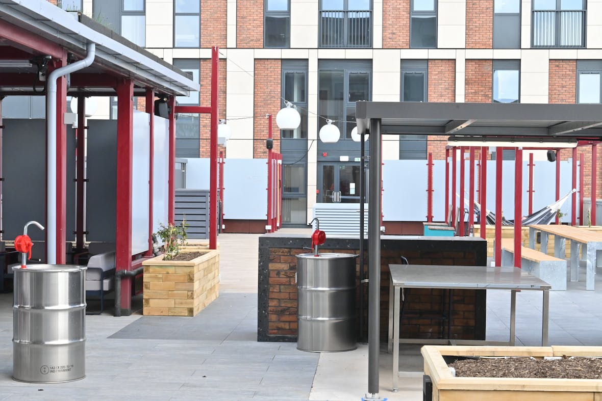 Modern outdoor terrace with red metal framework, stainless steel cylindrical planters, wooden raised beds, and brick apartment building in background.