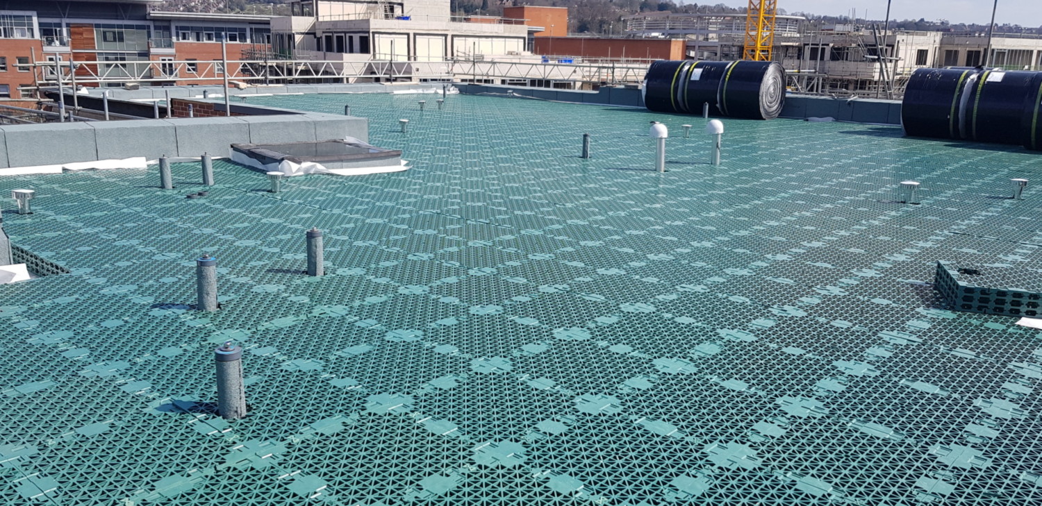 Green mesh decking covers rooftop surface with metal posts and cable reels visible, industrial buildings in background.