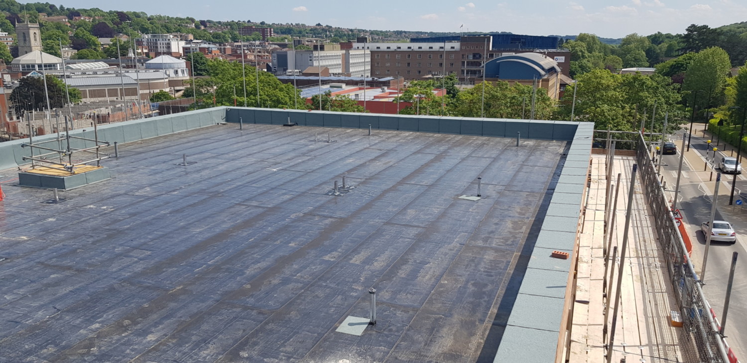 Large flat roof with dark grey membrane surface, white building edge, town buildings and green trees in background.