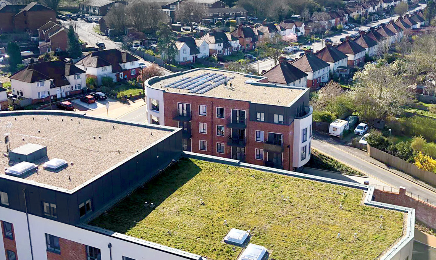 Aerial view of red brick apartment building with curved roof amongst residential houses, featuring green roof and suburban neighbourhood.