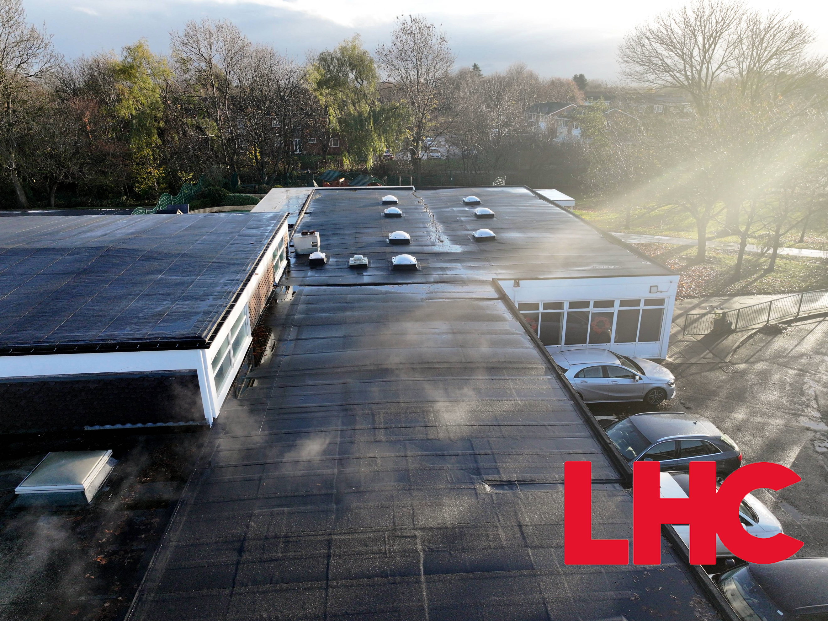 Aerial view of flat-roofed building with dark roofing, white walls, circular roof features, car park, and hills in background.
