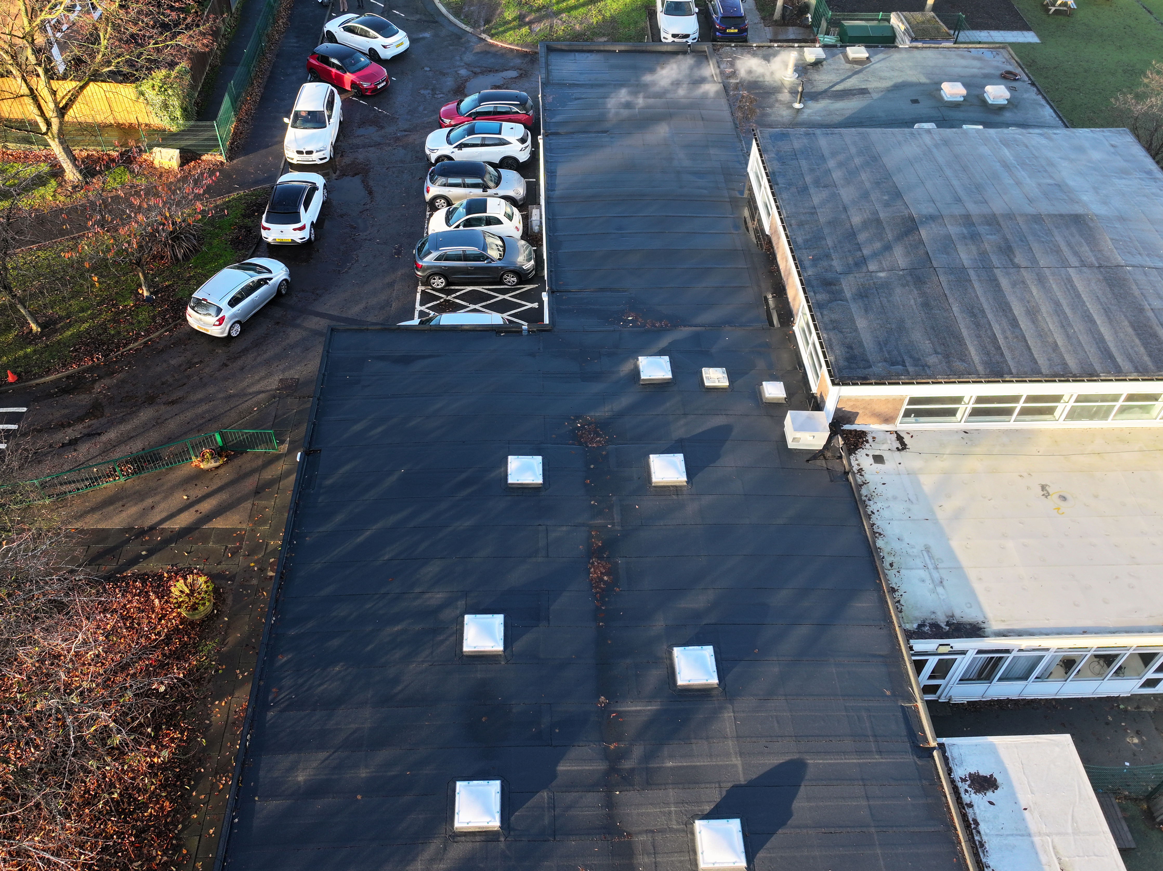 Aerial view of dark flat roof with white square vents, parked cars in adjacent car park, and surrounding green grass areas.