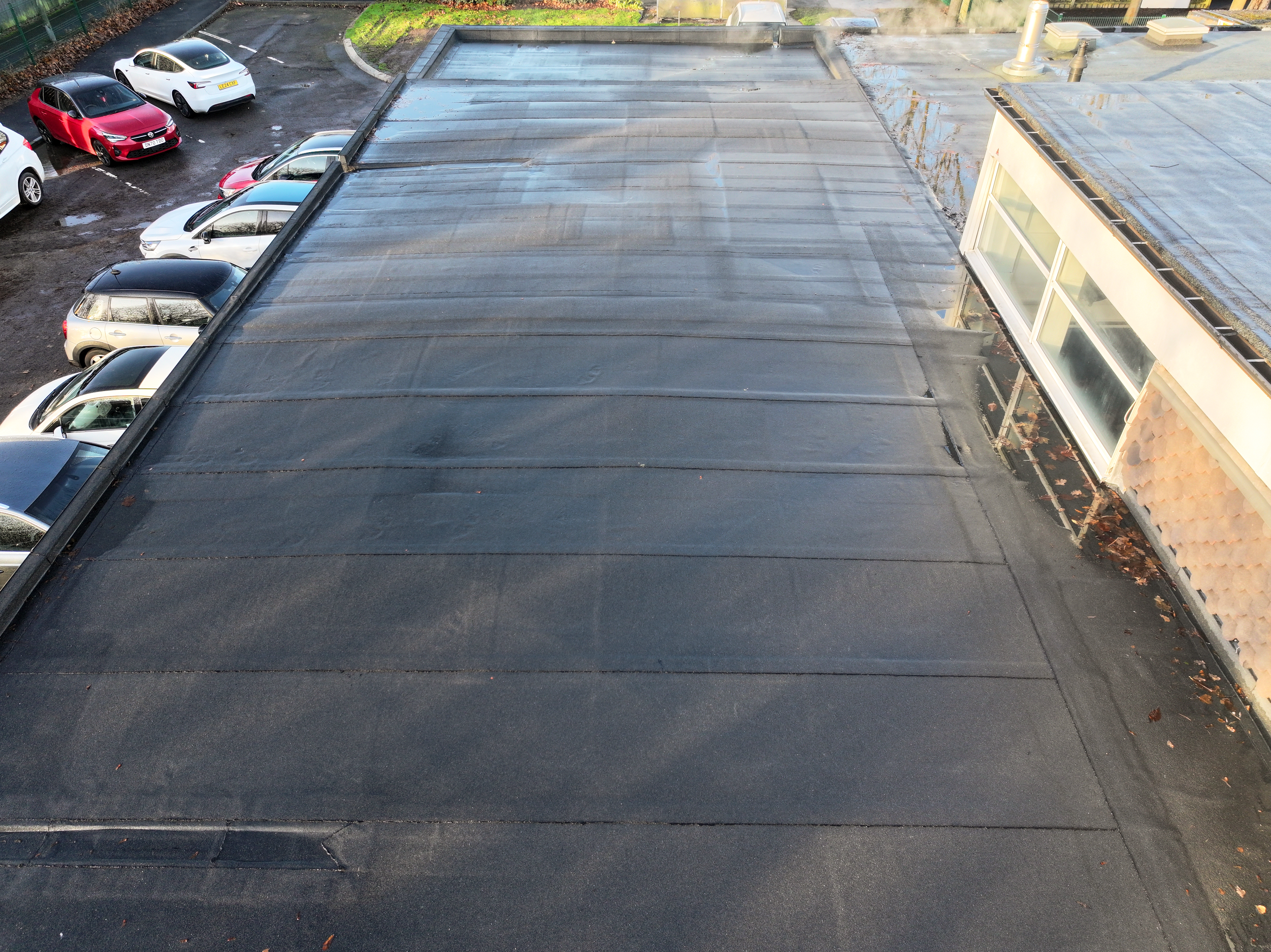 Aerial view of large concrete car park with grid pattern markings, parked cars along left edge, and cream-coloured building on right.