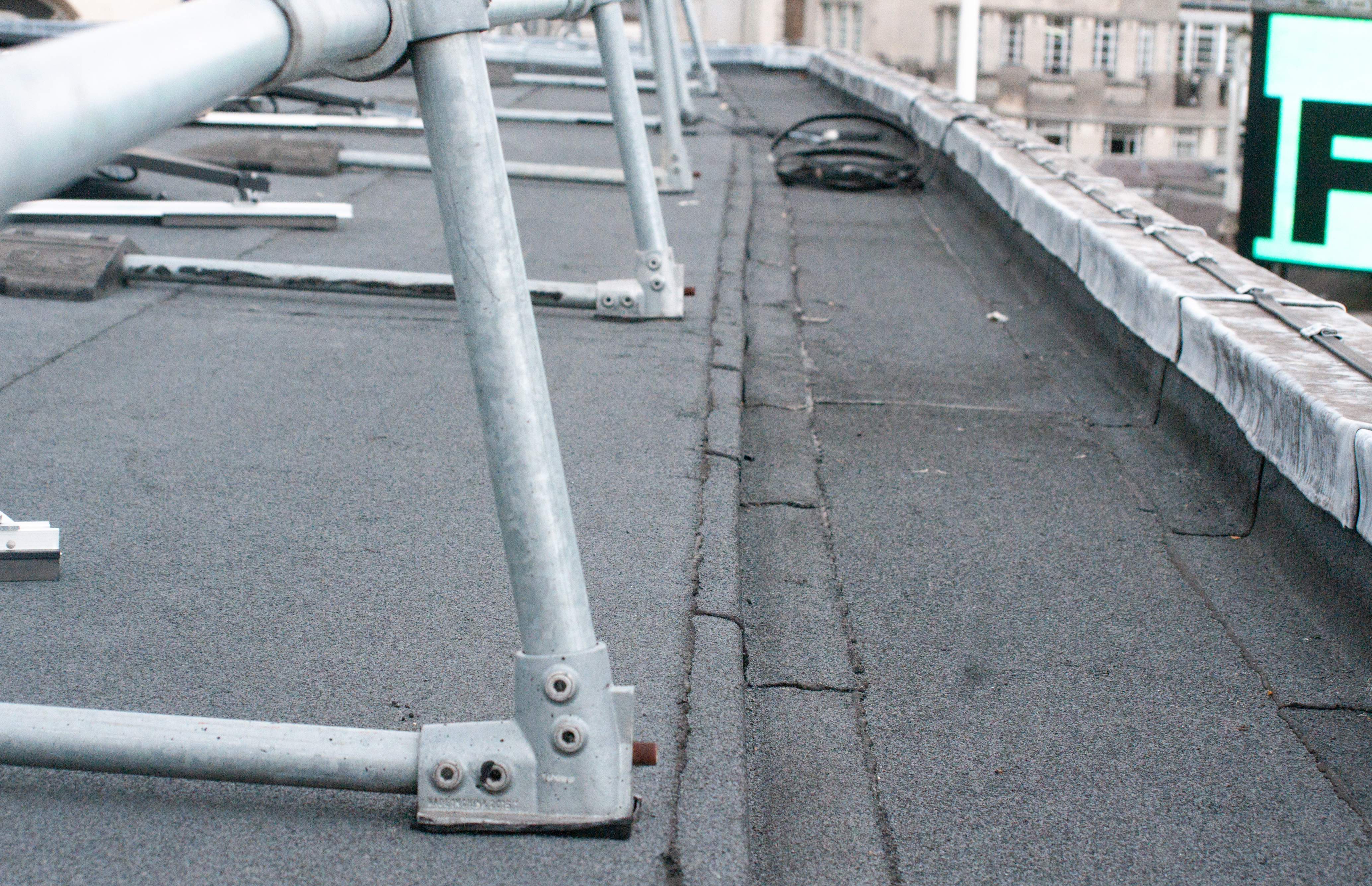 Grey rooftop with metal railings and support structures, cracked surface, beige building visible in background.