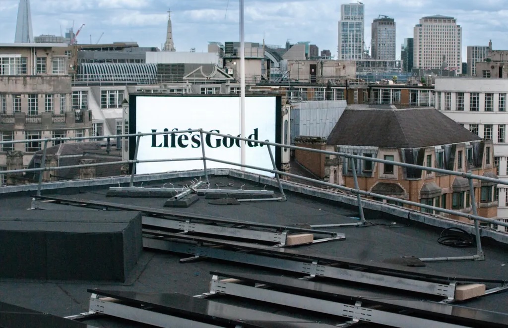 White billboard displaying "Life's Good." text on dark rooftop with city skyline in background under cloudy sky.