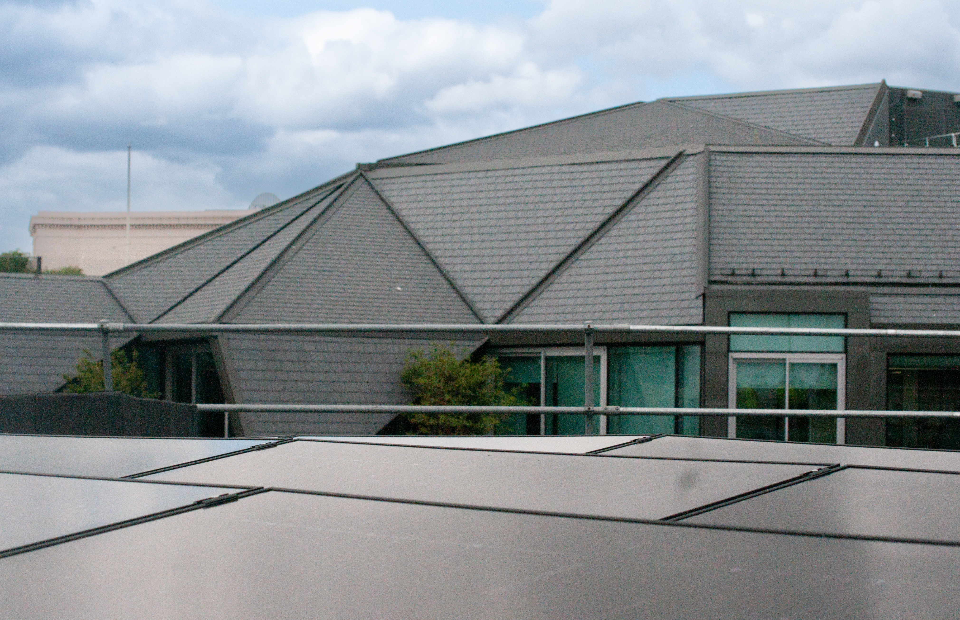 Modern building with angular grey metal roof, green window frames, and geometric paved forecourt under cloudy sky.
