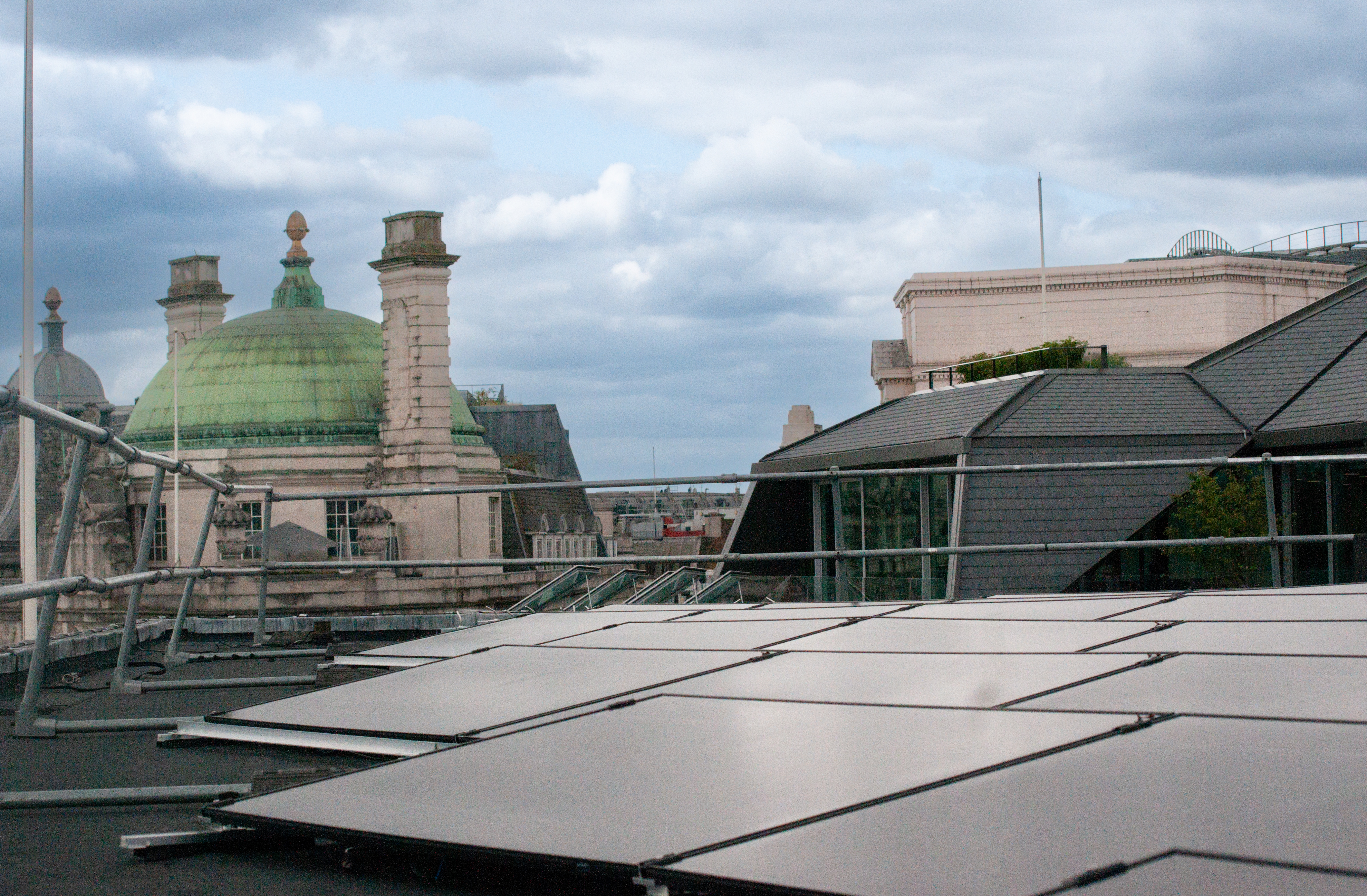 Solar panels on rooftop with various buildings and chimneys in background under cloudy sky.