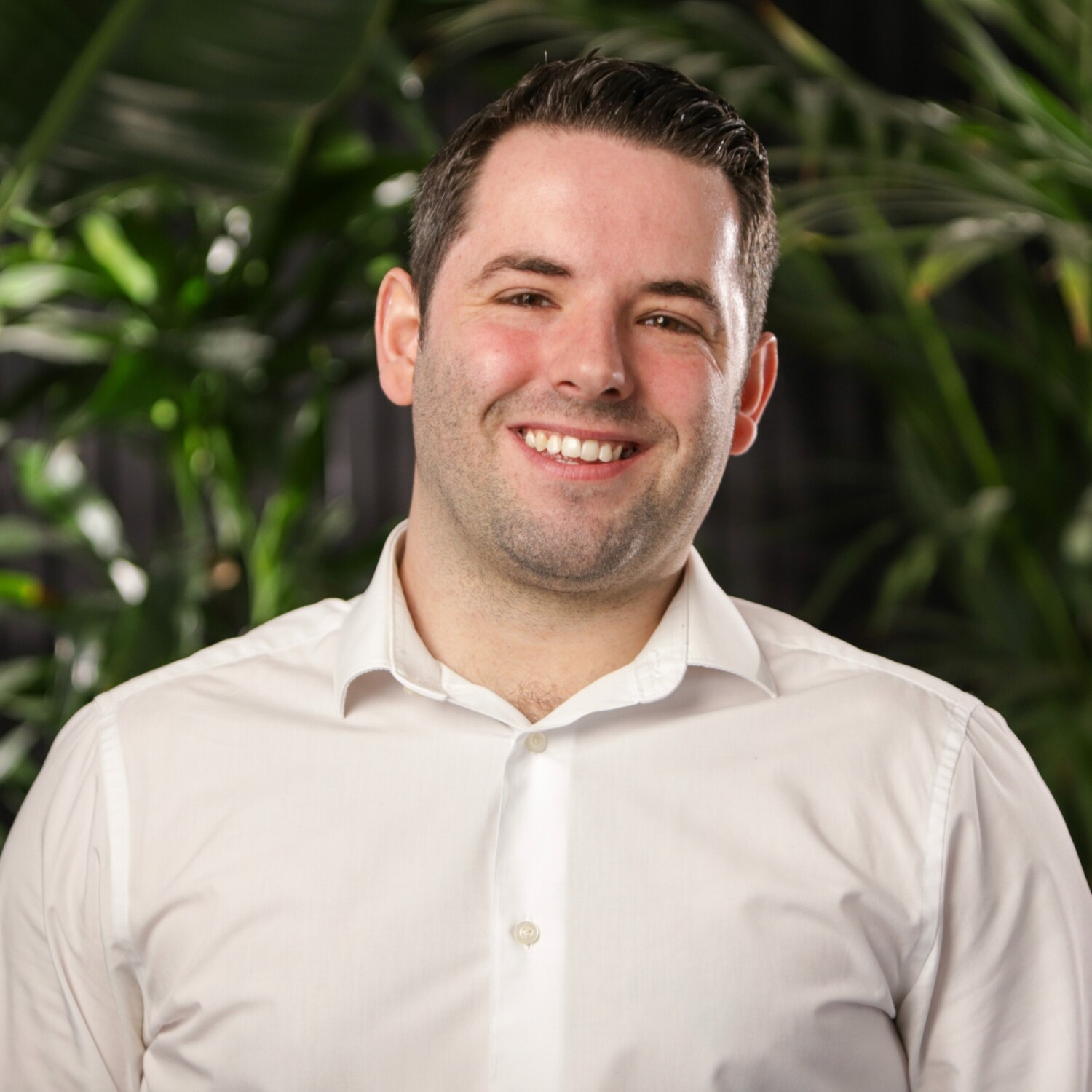 Man with short dark hair and facial stubble smiling, wearing white collared shirt against blurred green foliage background.