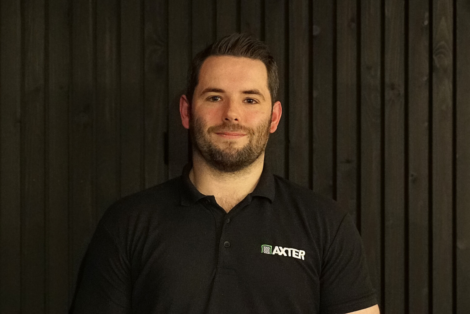 Man with beard wearing dark green polo shirt with "AXTER" logo, standing against dark wooden slat background.