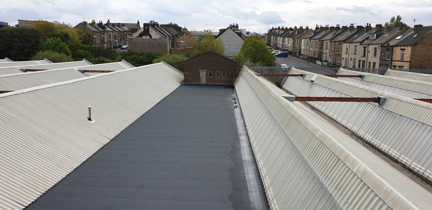 Aerial view of flat grey rooftop with white corrugated metal roofing on either side, residential houses visible in background under cloudy sky.