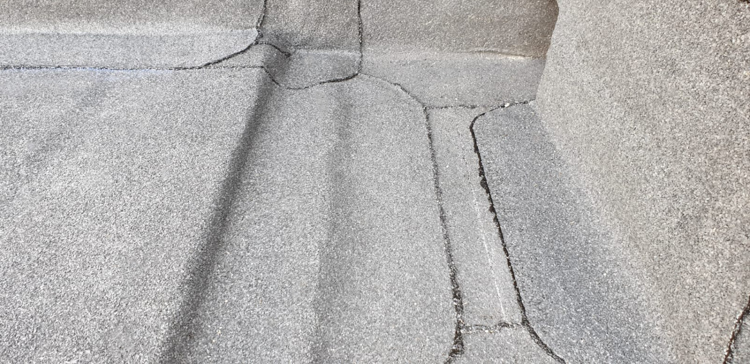 Cracked concrete pavement with diagonal shadow cast across grey surface, showing multiple fracture lines and weathered texture.