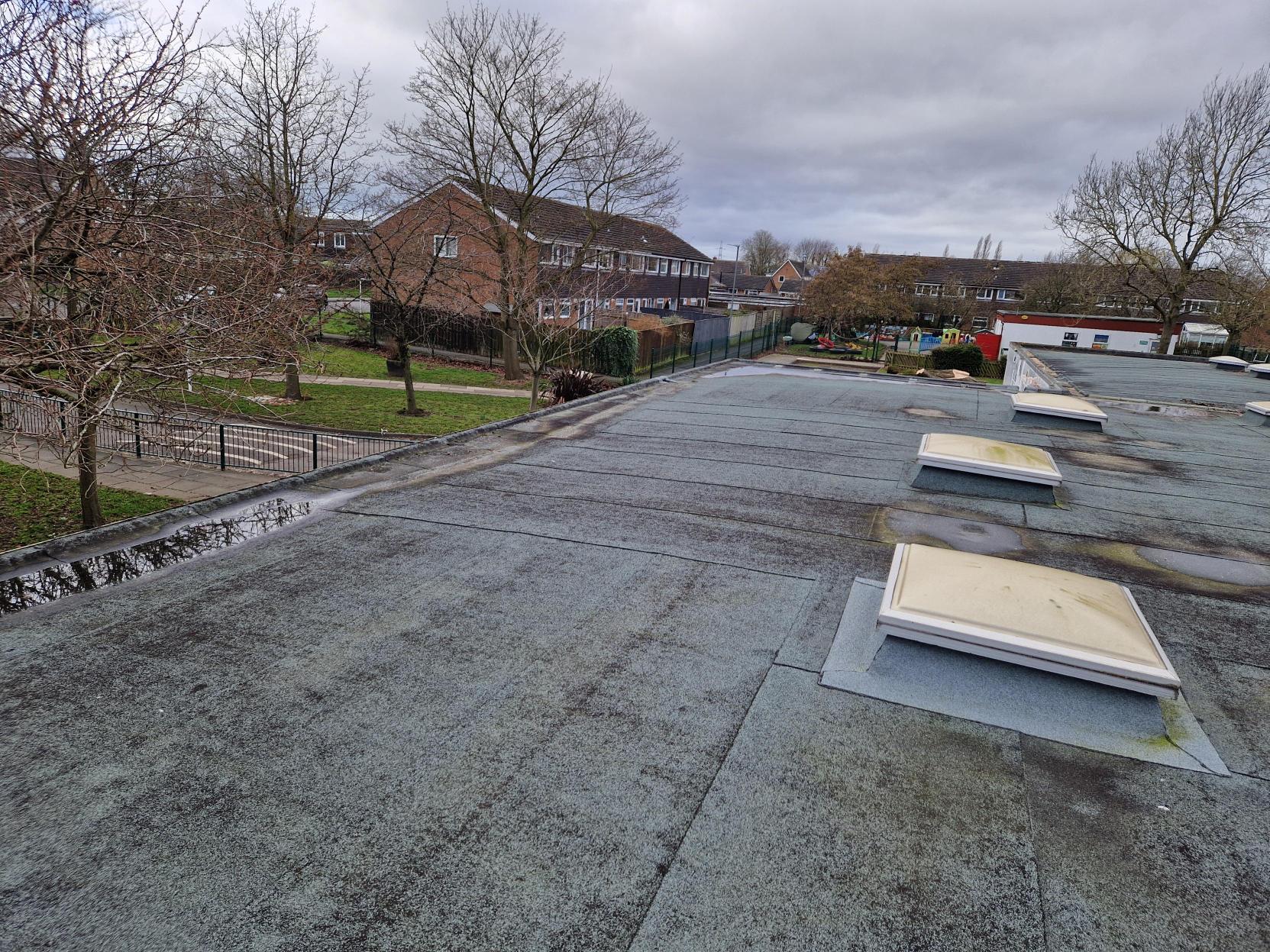 Flat roof with rectangular white vents, overlooking residential area with brick houses, bare trees, and cloudy grey sky.