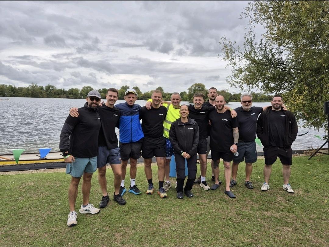 Group of rowers in dark athletic wear posing on grass beside lake, with rowing boats visible in background under cloudy sky.