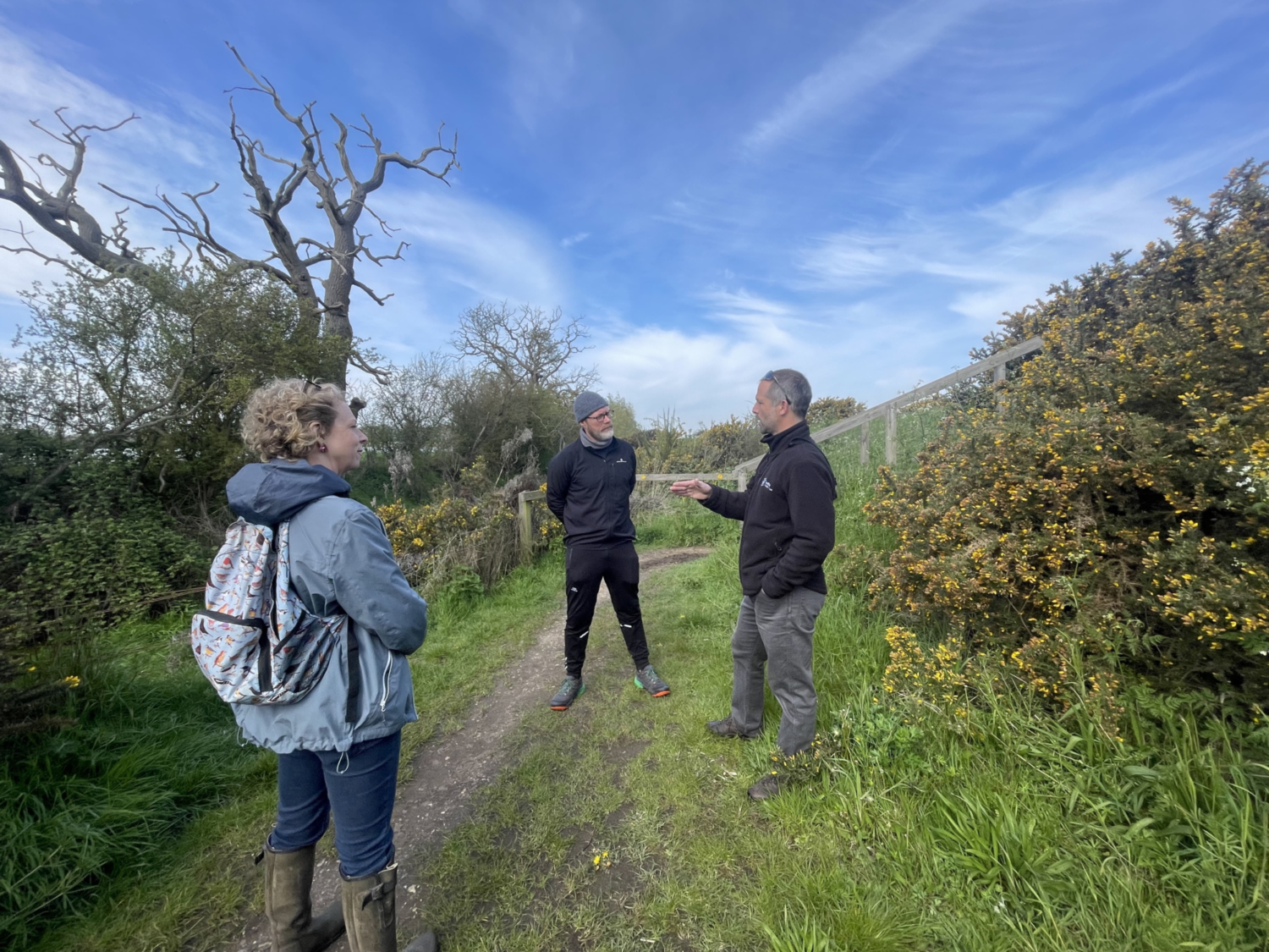 Three people standing on dirt path surrounded by green hedgerows and bare trees under blue sky with white clouds.