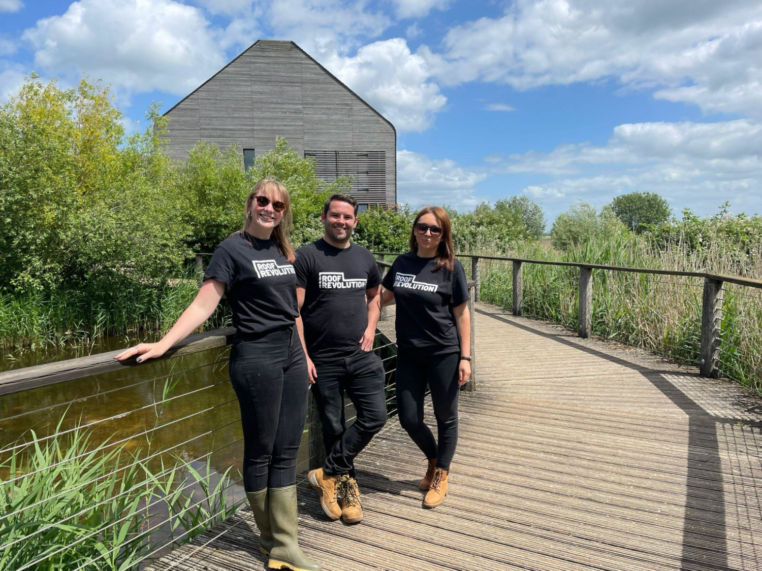 Three people in black t-shirts standing on wooden boardwalk with metal railings. Grey wooden building and green trees in background under blue sky with white clouds.