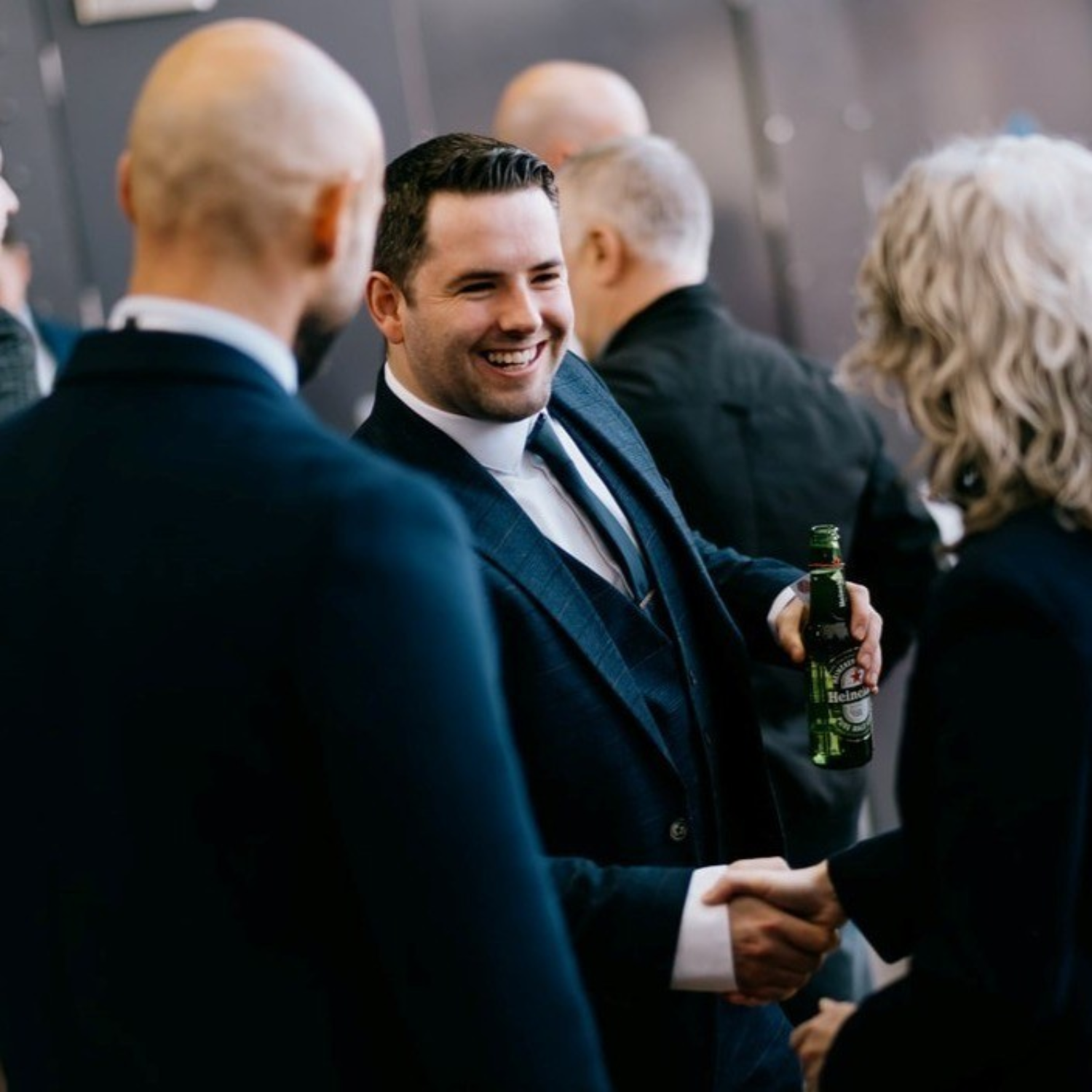 Man in dark suit smiling whilst holding green bottle, surrounded by other formally dressed people at social gathering.