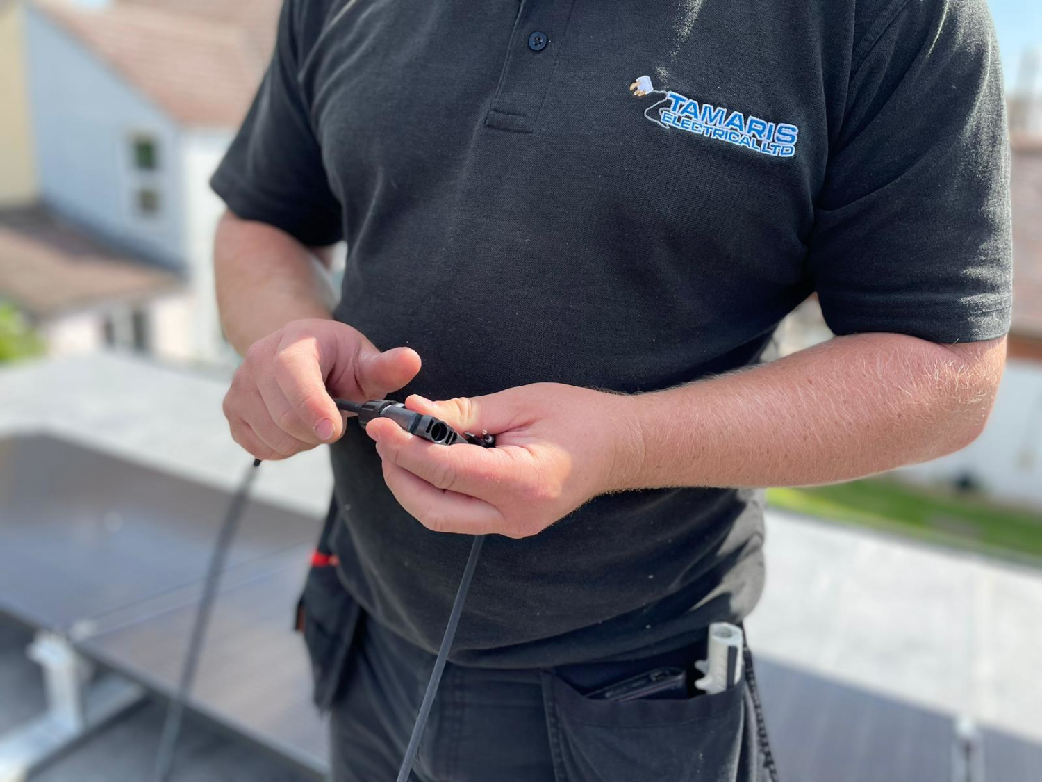 Man in dark polo shirt with blue logo holding black cable or wire, residential street background visible.